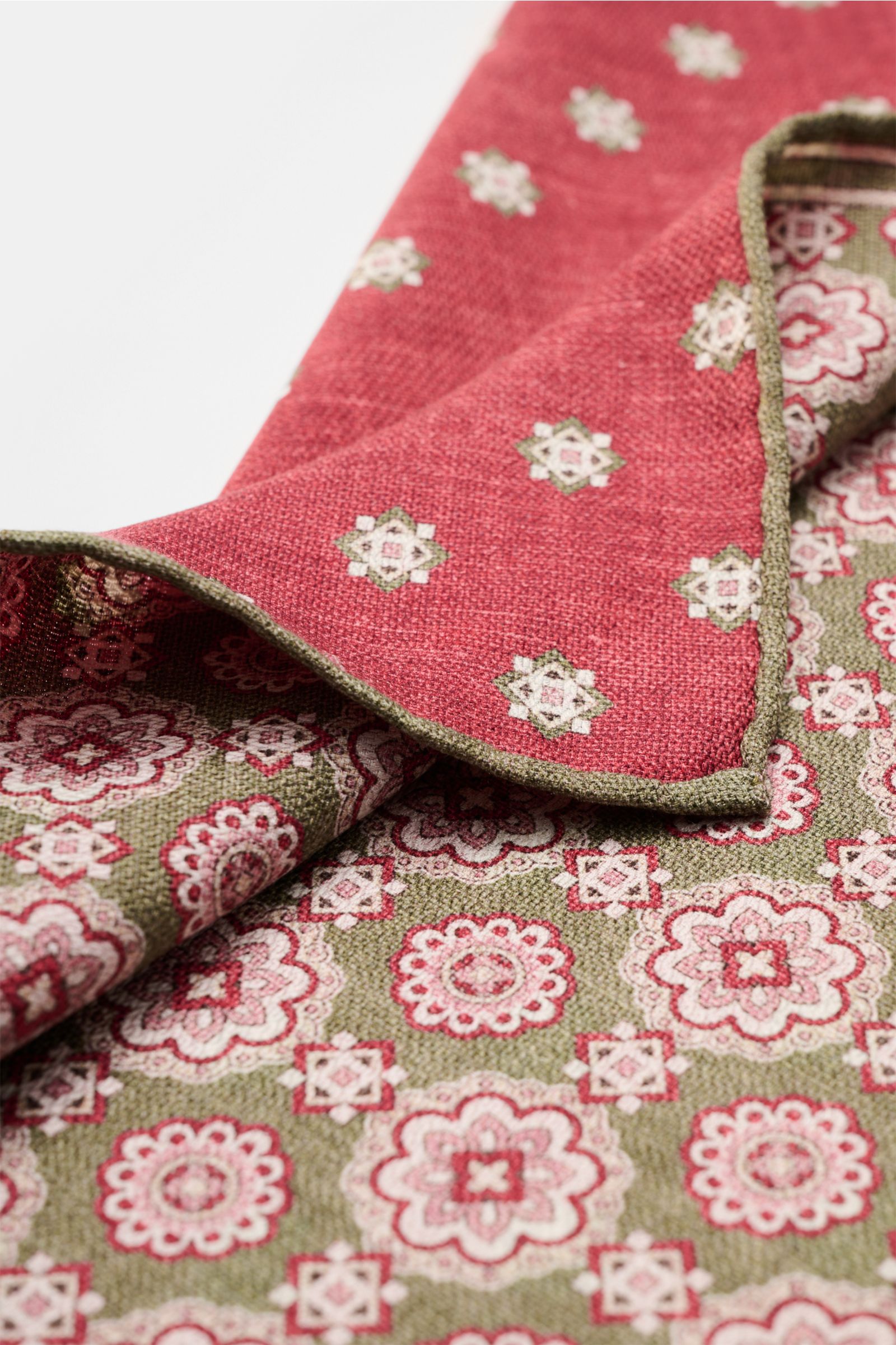Close-up photo from a side angle of the Rosi Collection Seiden-Einstecktuch 'Easy' oliv/rot/rosé gemustert showing elegant medallion print with border, contrasting reverse side, pure silk, soft smooth feel, rolled edges, Made in Italy, approx. 31 cm x 31 