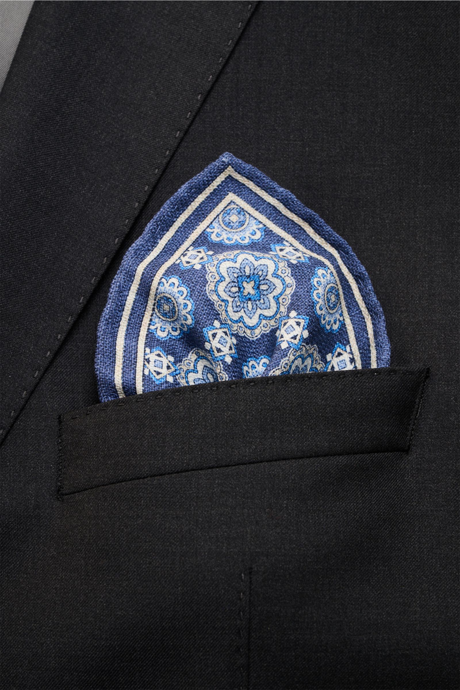 Close-up front view of Rosi Collection Seiden-Einstecktuch 'Easy' dunkelblau/blau/creme gemustert in a black suit pocket, reine Seide, softer, geschmeidiger Griff, eleganter Medaillon-Print mit Borte, kontrastierende Rückseite, rollierte Kanten, Made in I