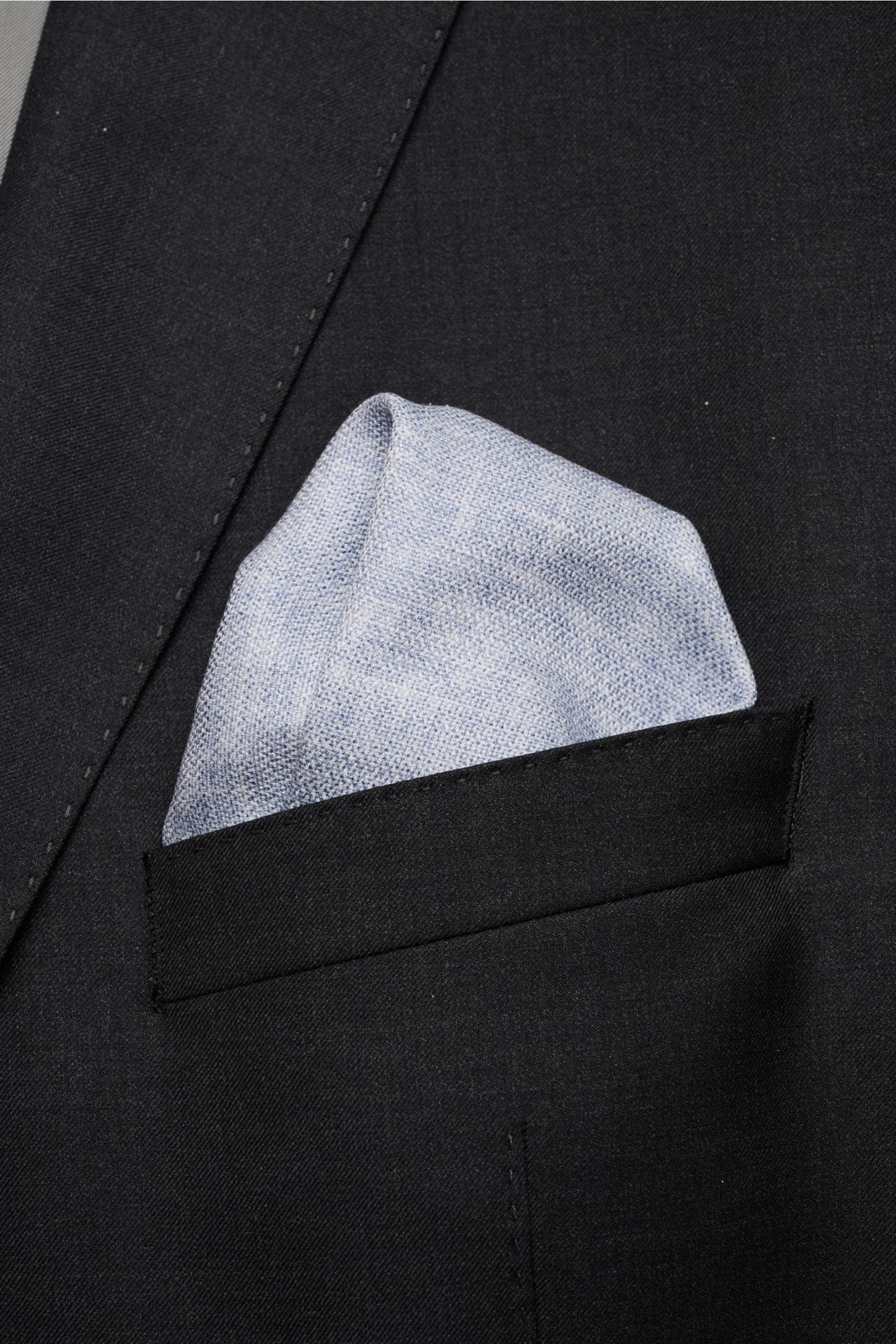 Brunello Cucinelli Seiden-Einstecktuch rauchblau meliert folded in the breast pocket of a black suit jacket, photographed from above. Quiet Luxury kann BRUNELLO CUCINELLI besonders gut und bringt auch in dieser Saison passende Einstecktücher, wie diese