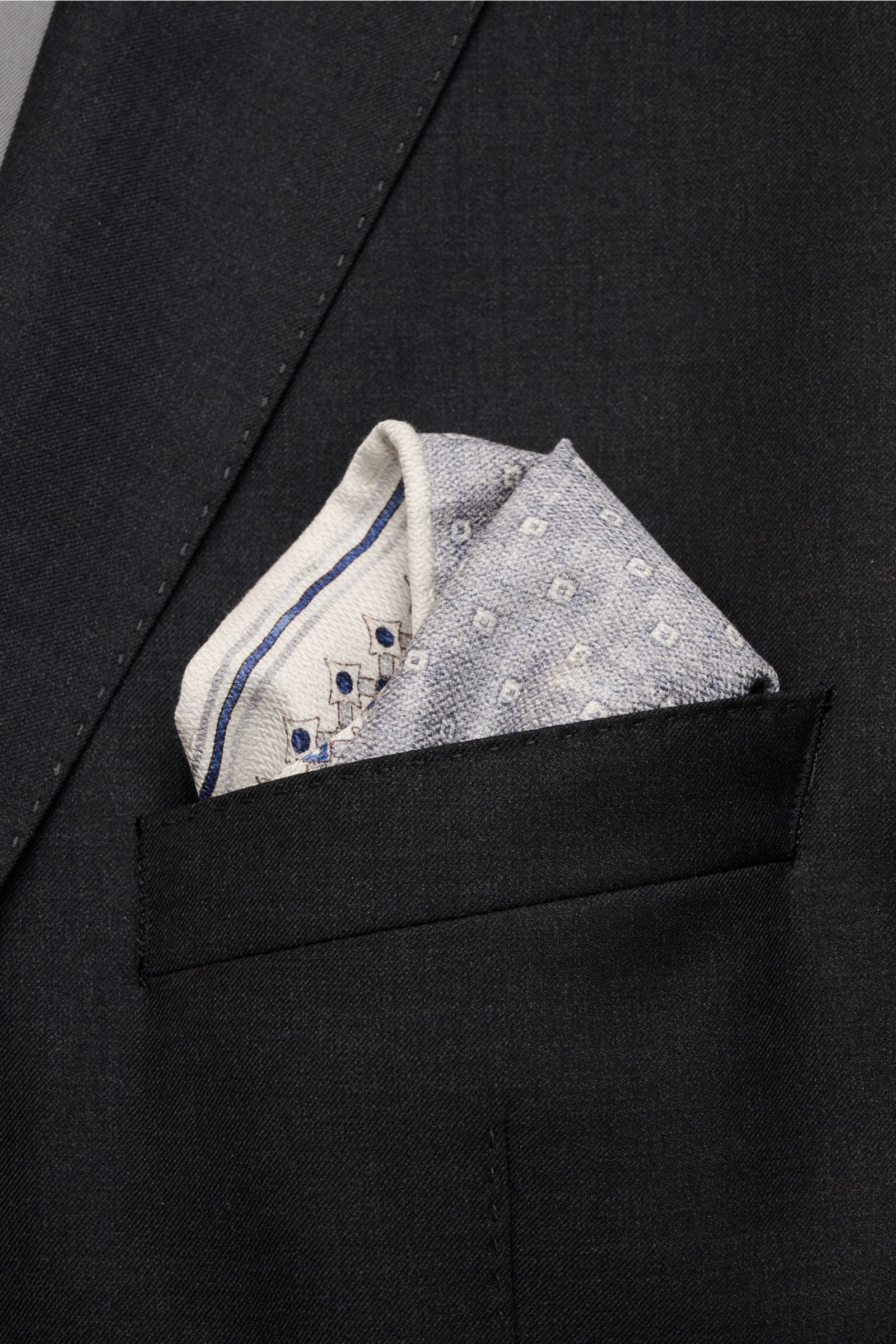 Nahaufnahme eines schwarz-grauen Sakkos mit einem aus der Brusttasche herausragenden, sand/navy/grau gemusterten Brunello Cucinelli Seiden-Einstecktuch mit grafischem Print, rollierten Kanten und weichem Griff, Vorder- und Rückseite unterschiedlich gemust