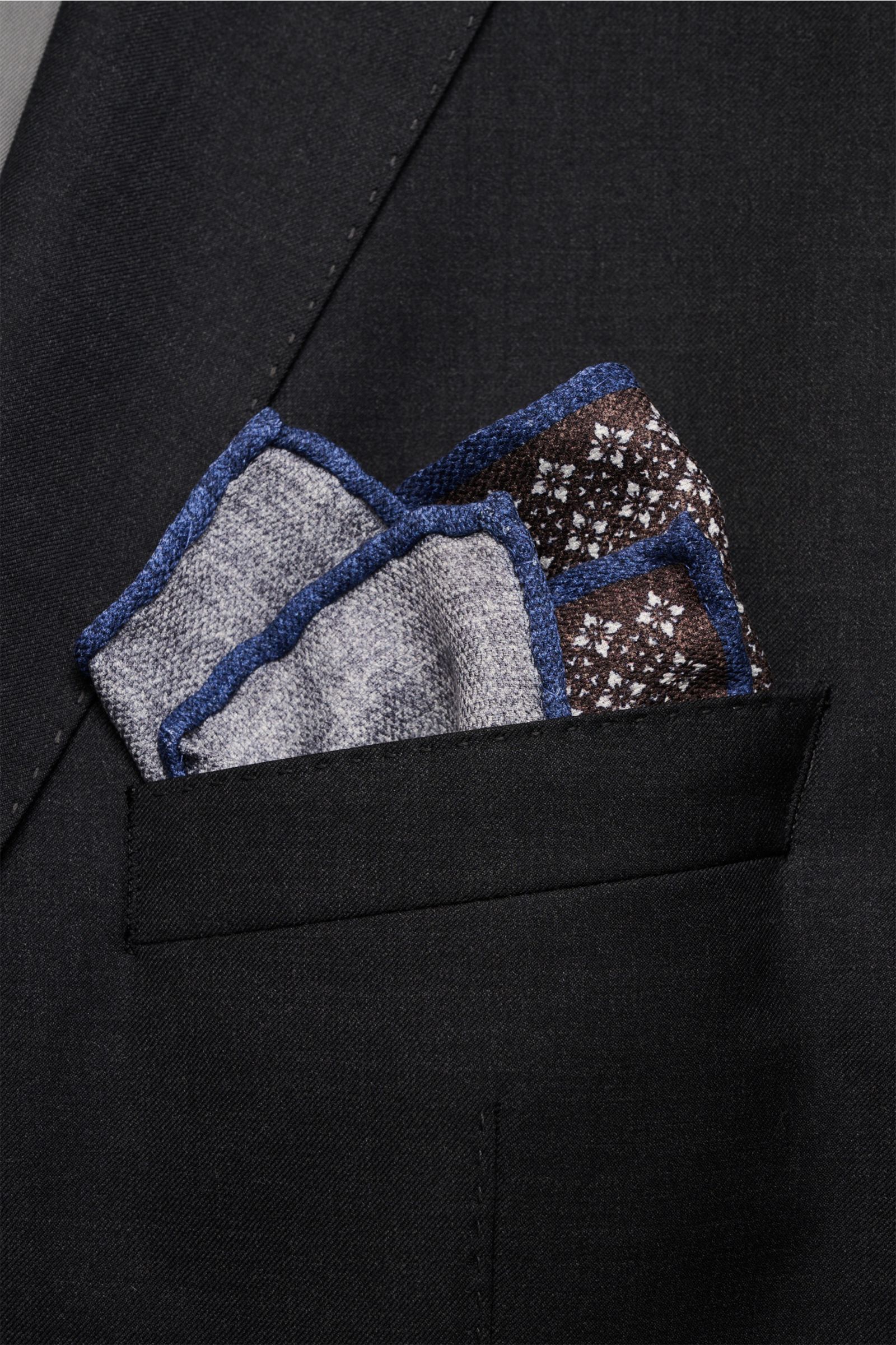 Brunello Cucinelli Seiden-Einstecktuch dunkelbraun/navy/sand gemustert folded in a black suit pocket, photographed from above; Quiet Luxury heißt der große Trend: Das kann BRUNELLO CUCINELLI besonders gut und bringt auch in dieser Saison passende Einsteck