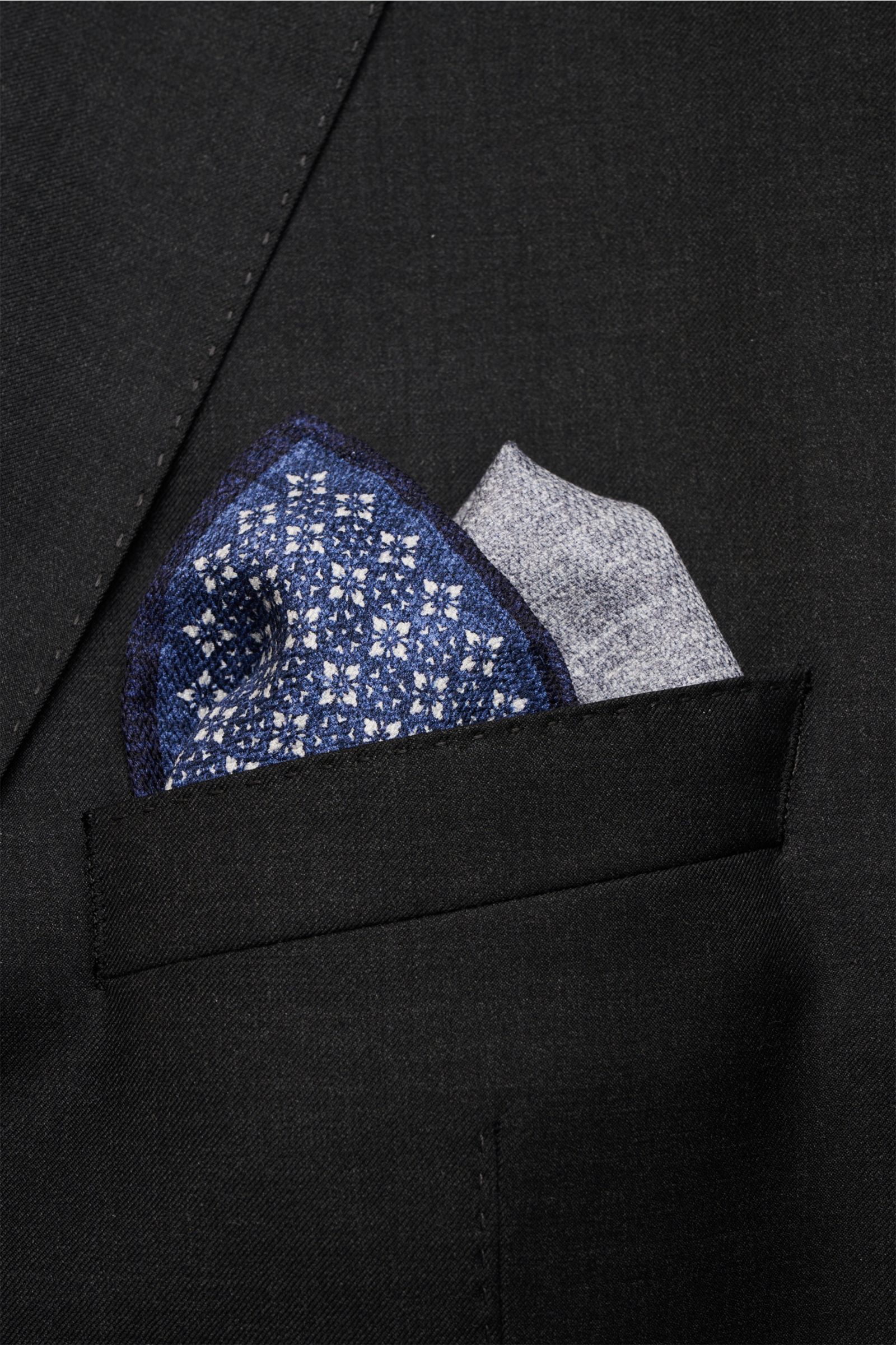 Close-up front view of a Brunello Cucinelli Seiden-Einstecktuch dunkelblau/grau gemustert pocket square in a black jacket pocket, showcasing its floral pattern and hand-rolled edges.

Description: Quiet Luxury heißt der große Trend: Das kann BRUNELLO C