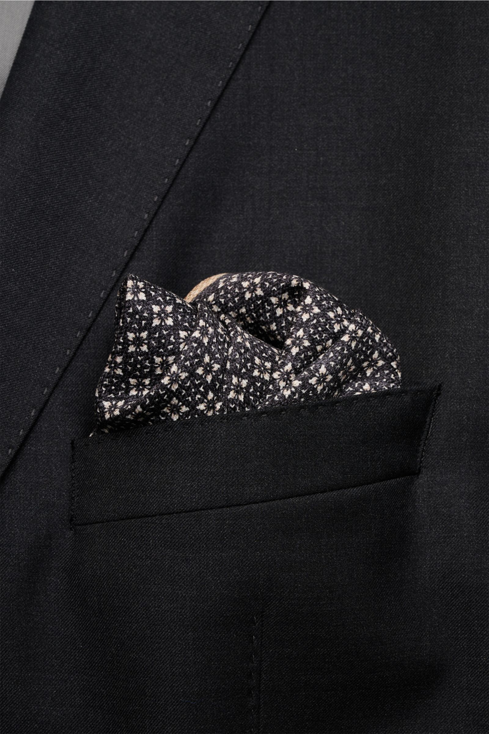 Close-up front view of a black suit jacket pocket featuring a floral patterned Brunello Cucinelli Seiden-Einstecktuch anthrazit/graubraun/sand gemustert made of pure silk. Quiet Luxury heißt der große Trend: Das kann BRUNELLO CUCINELLI besonders gut und b