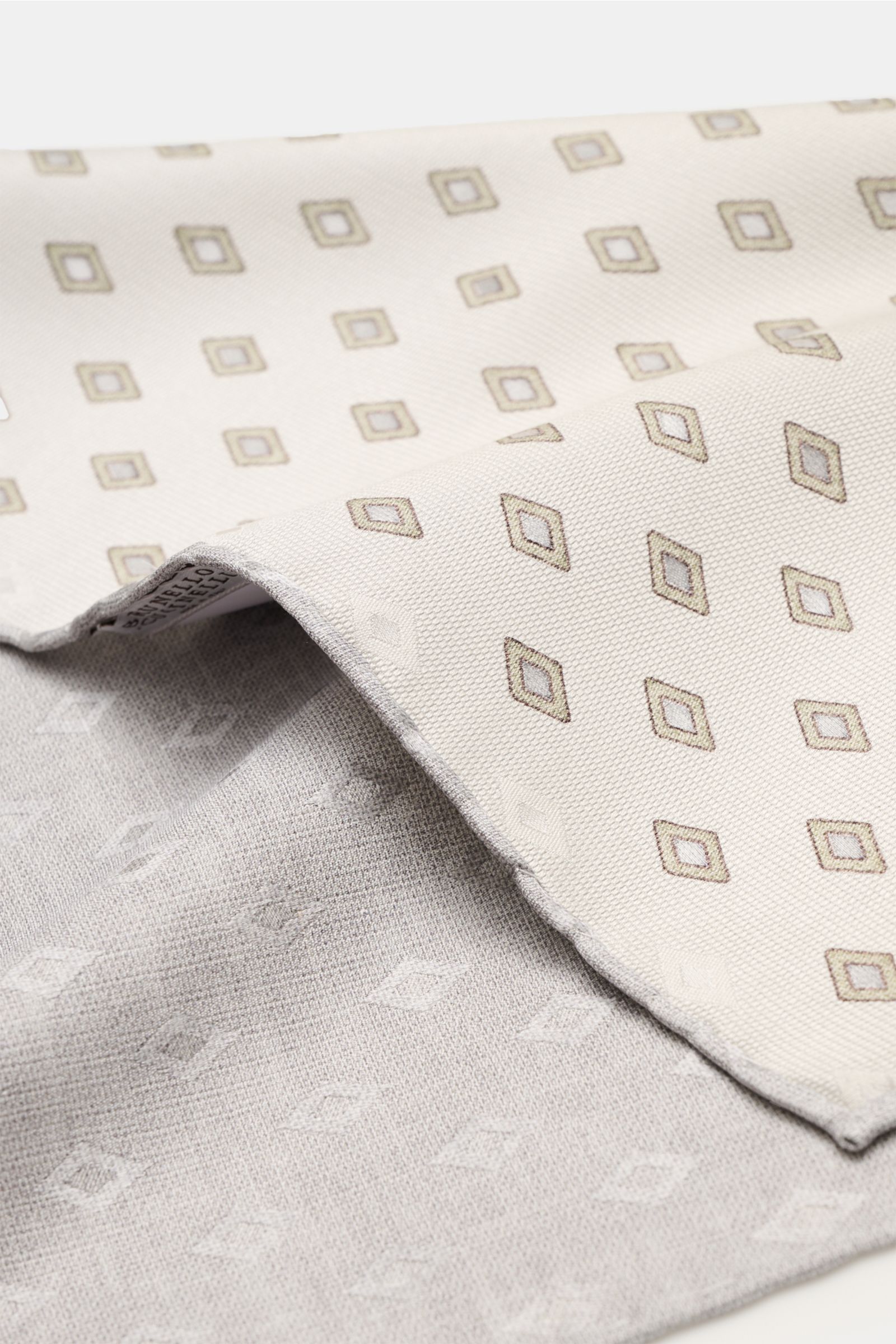 Close-up from an angled top view of the Brunello Cucinelli Seiden-Einstecktuch sand/salbei/grau gemustert, showing its elegant rhombus pattern on pure silk with smooth texture, contrasting backside, and finely rolled edges in a contrasting color. Elega