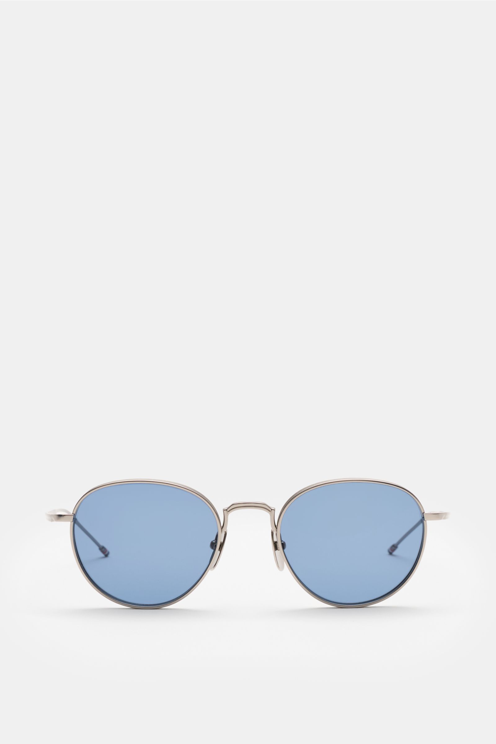 Front view of Thom Browne Sunglasses silver-grey with rounded shape, evenly tinted blue plastic lenses, deep metal frame, tricolour temple detail, and 100% UV protection; hard case included.