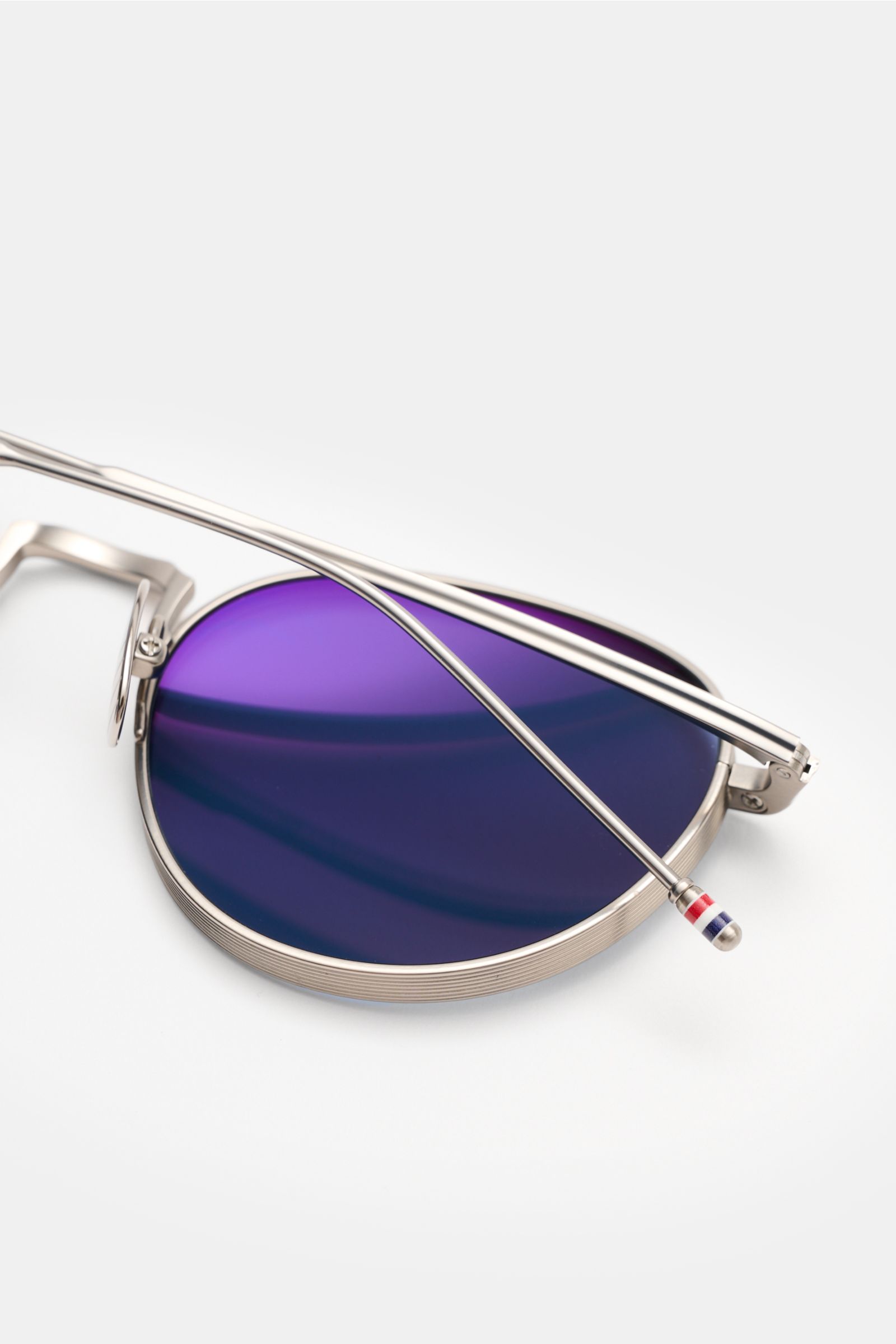 Thom Browne Sunglasses silver-grey shown from a close-up side angle, featuring a rounded shape, deep metal frame, evenly tinted purple lenses with 100% UV protection, and tricolour detail on the temple.