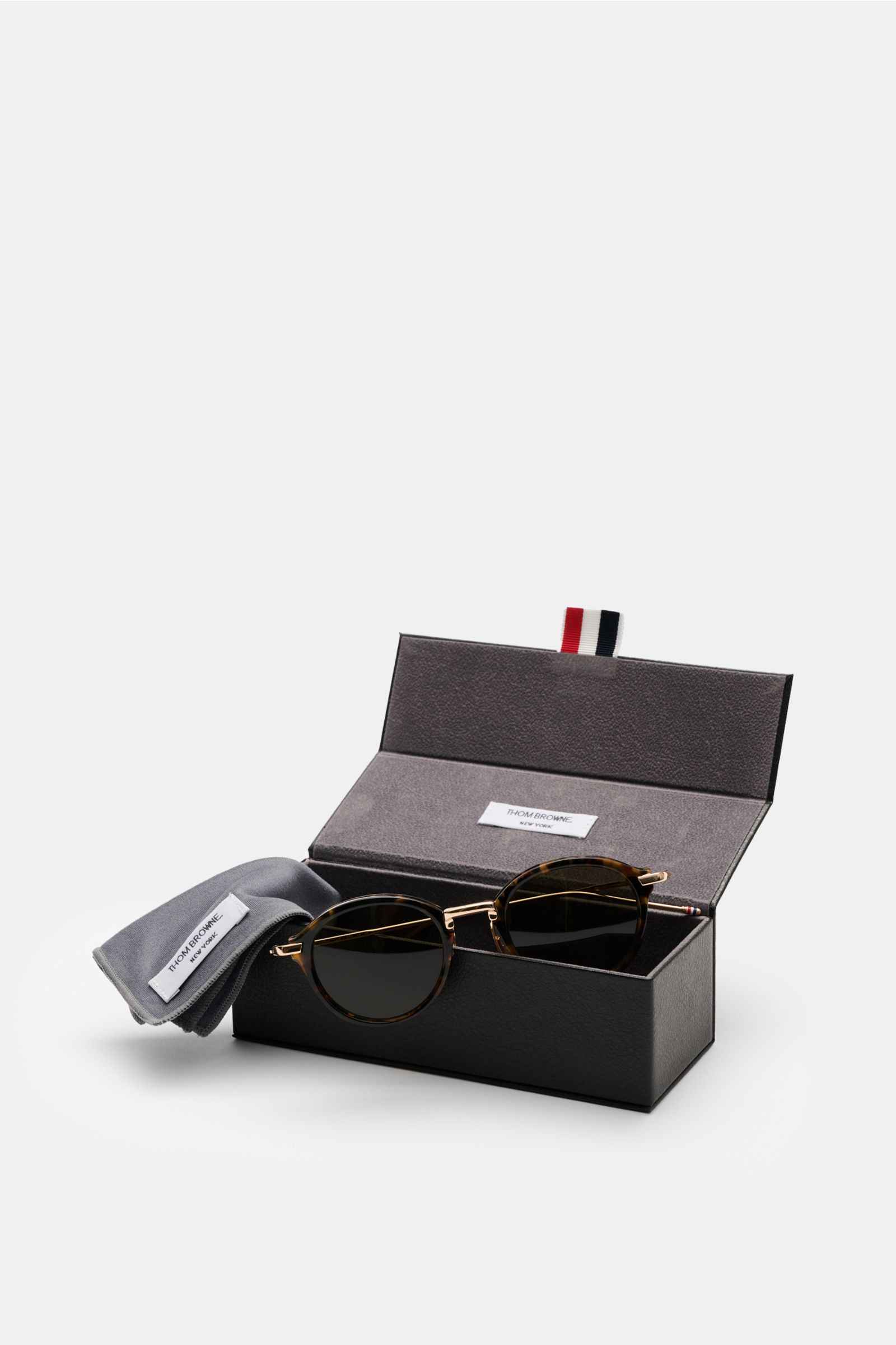 Thom Browne Sunglasses dark brown/gold patterned with round tortoiseshell acetate frame and evenly tinted lenses, shown front-left in open black case with grey cleaning cloth.

These round sunglasses by THOM BROWNE have a tortoiseshell design and tricolou