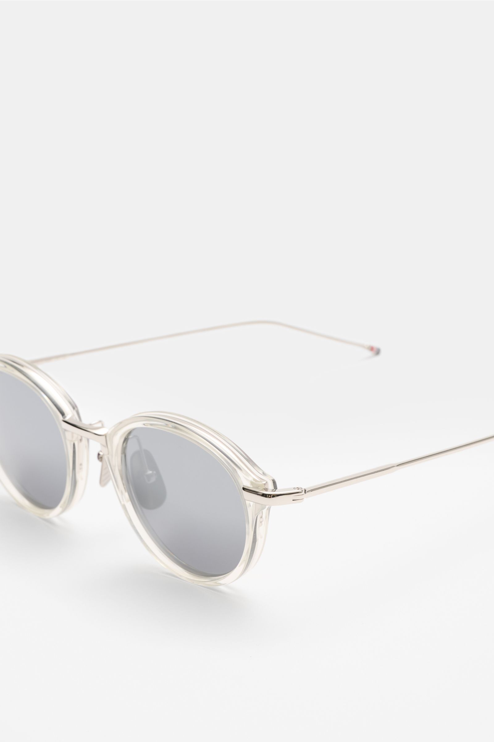 Nahaufnahme der Thom Browne Sonnenbrille hellgrau mit abgerundeter Form, transparentem Acetat-Rahmen und Tricolore-Detail am Bügel, seitliche Perspektive.