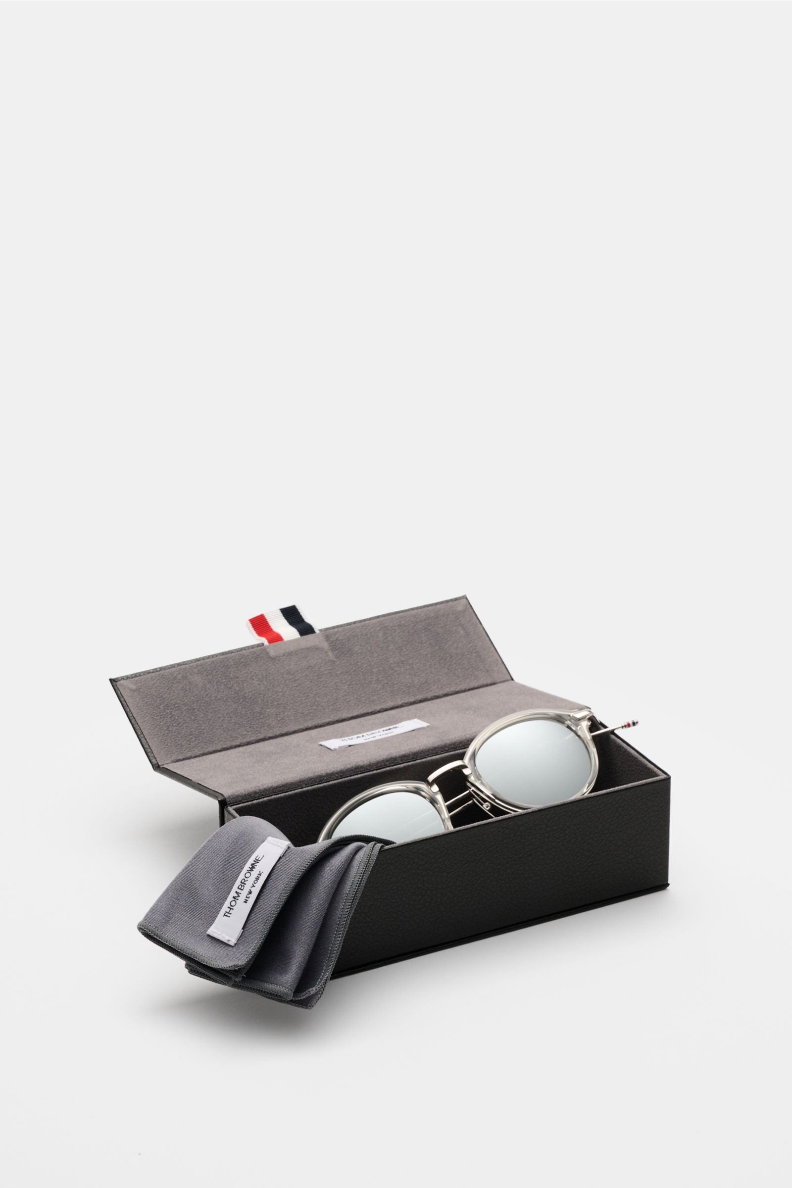 Thom Browne Sonnenbrille hellgrau mit abgerundeter Form, transparentem Acetat-Rahmen, getönten Gläsern, Tricolore-Detail im geöffneten Etui, seitliche Perspektive.