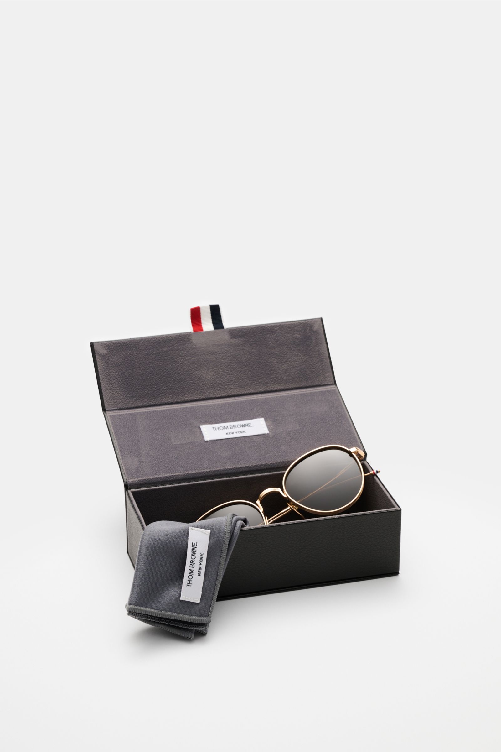 Thom Browne Sonnenbrille gold in abgerundeter Form mit tiefem Metallrahmen, Tricolore-Detail, 100% UV-Schutz, leicht getönten Gläsern, im geöffneten Etui.
