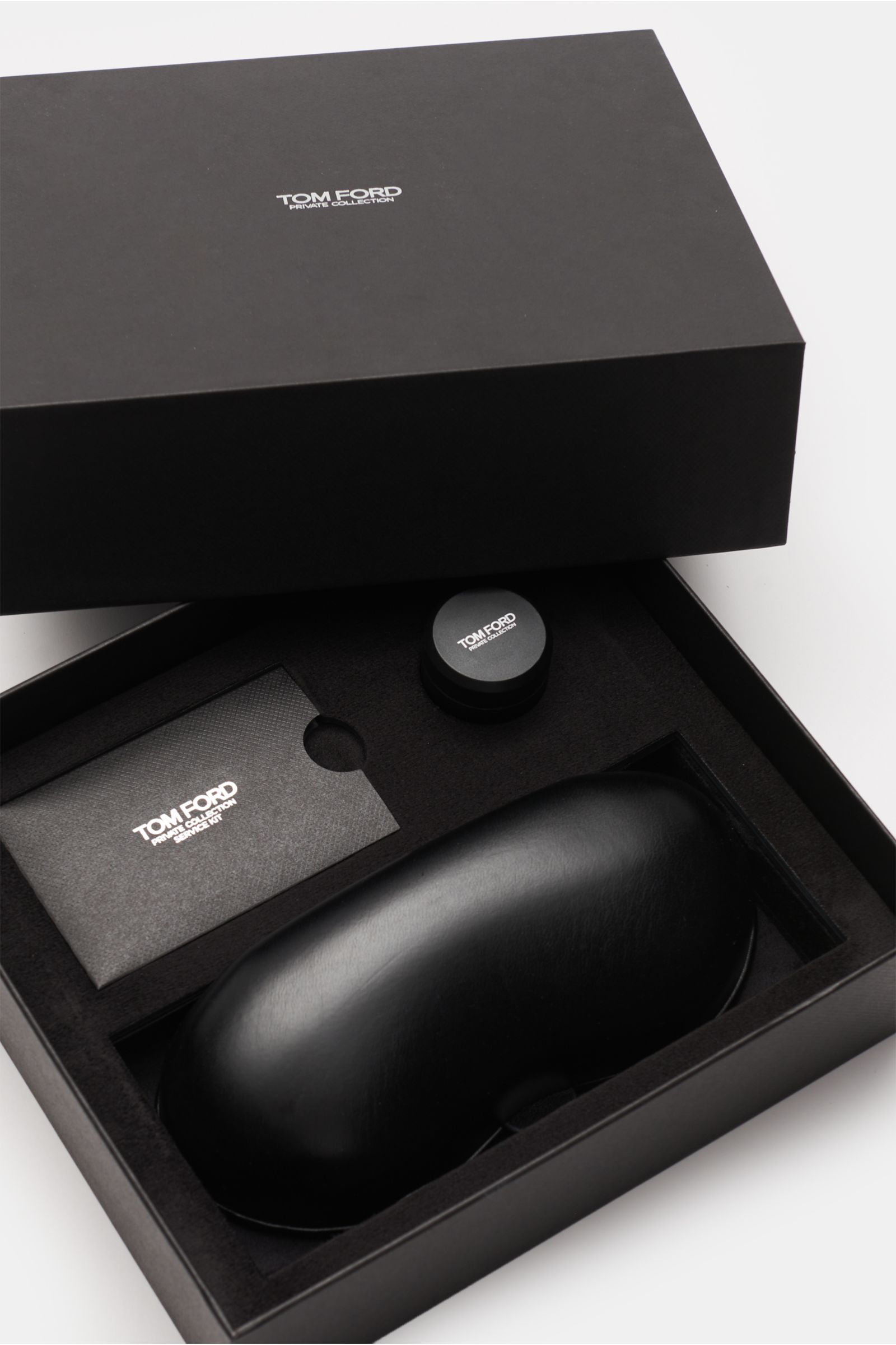 Geöffnete schwarze Geschenkbox mit Tom Ford Horn-Sonnenbrille 'Private Collection' anthrazit gemustert/gelb im Leder-Hartschalenetui, Politurset und Servicekarte von oben fotografiert.