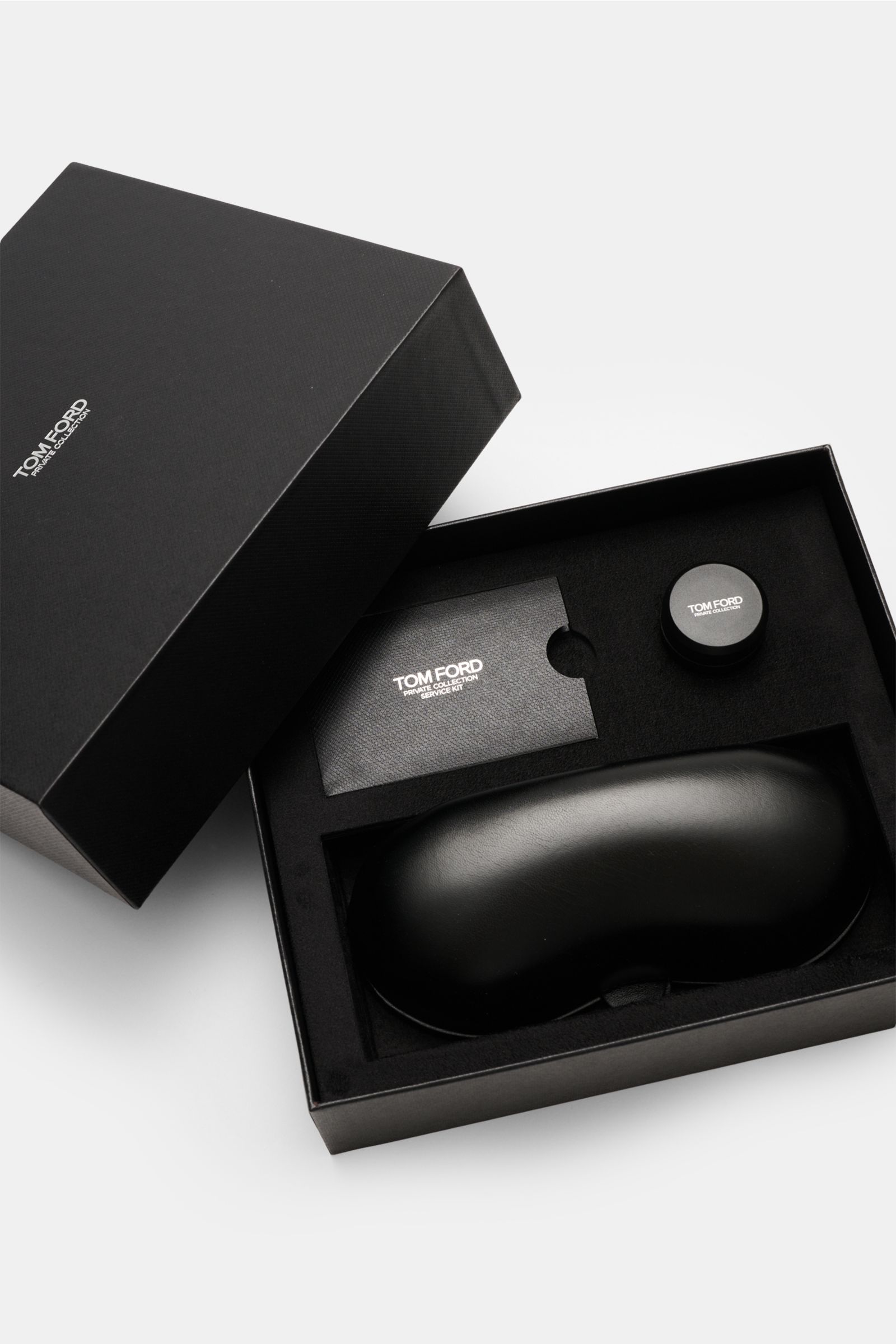 Das Bild zeigt aus seitlich-oben Perspektive die geöffnete schwarze Geschenkbox mit Leder-Hartschalenetui, Horn-Politurset und Putztuch der Tom Ford Horn Brille 'Private Collection' schwarz/grau gemustert, die runde Form, echten Büffelhornrahmen, blaulich