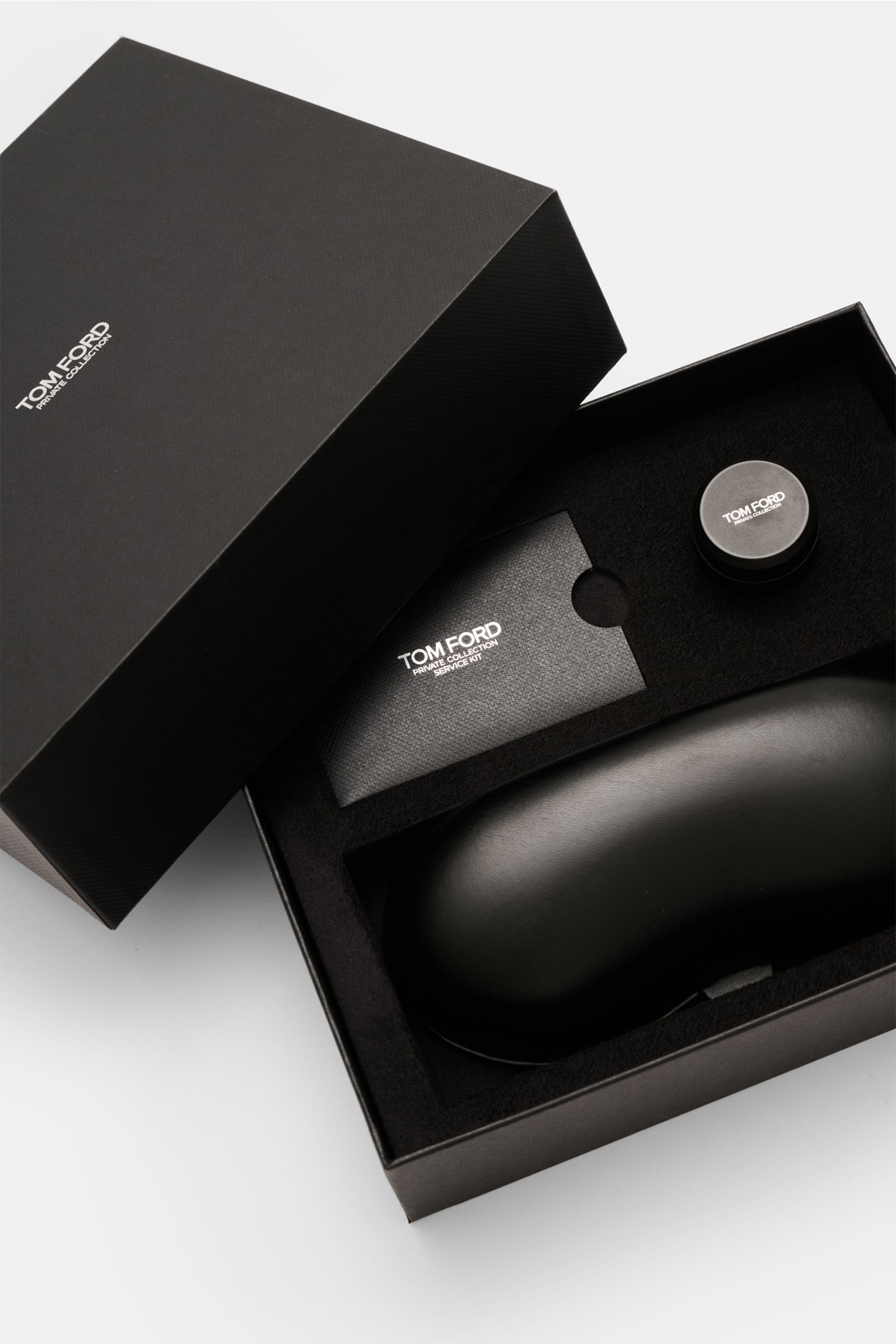 Das Bild zeigt aus der Vogelperspektive die geöffnete Verpackung der Tom Ford Horn Brille 'Private Collection' schwarz/grau gemustert mit Lederetui, Politurset und Putztuch.