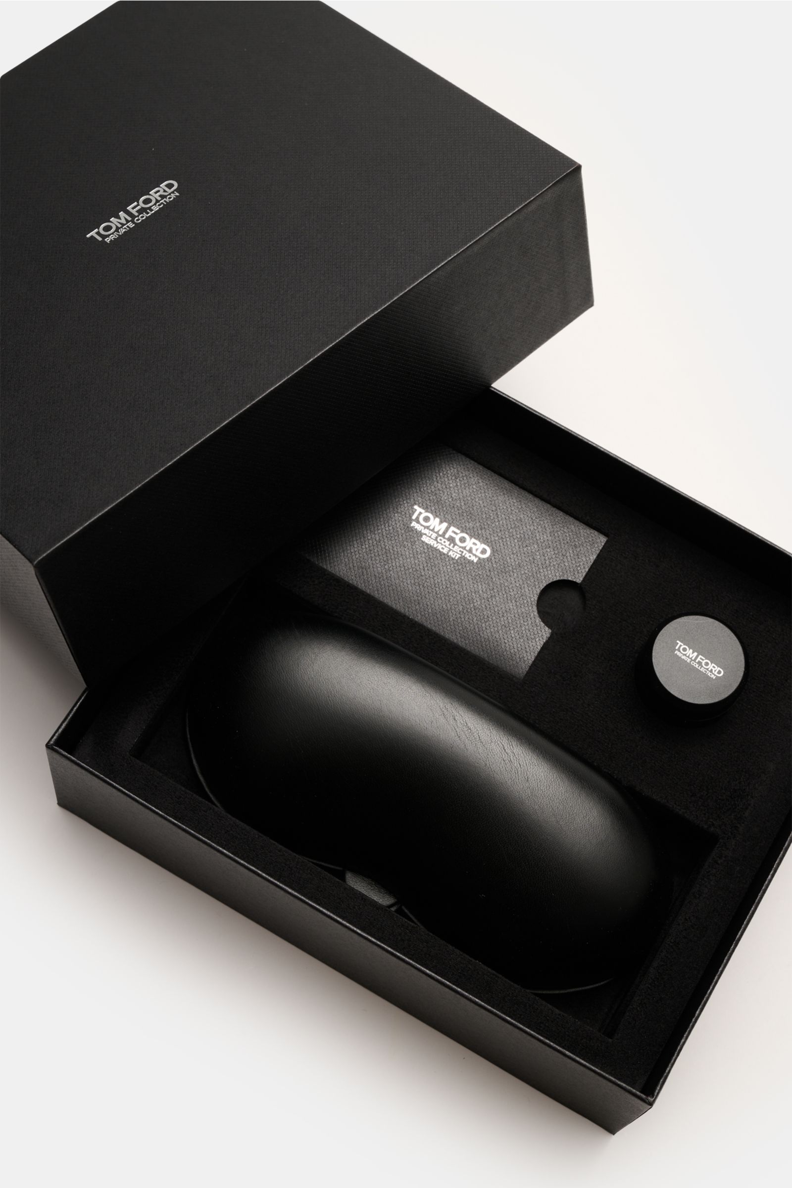 Opened black Tom Ford box viewed from above, showing a leather hard case, a small round horn polish container, and a card, all from the Tom Ford Horn Brille 'Private Collection' schwarz/graubraun/weiß gemustert set. 

Stilvoller Eyecatcher: Die exklusi