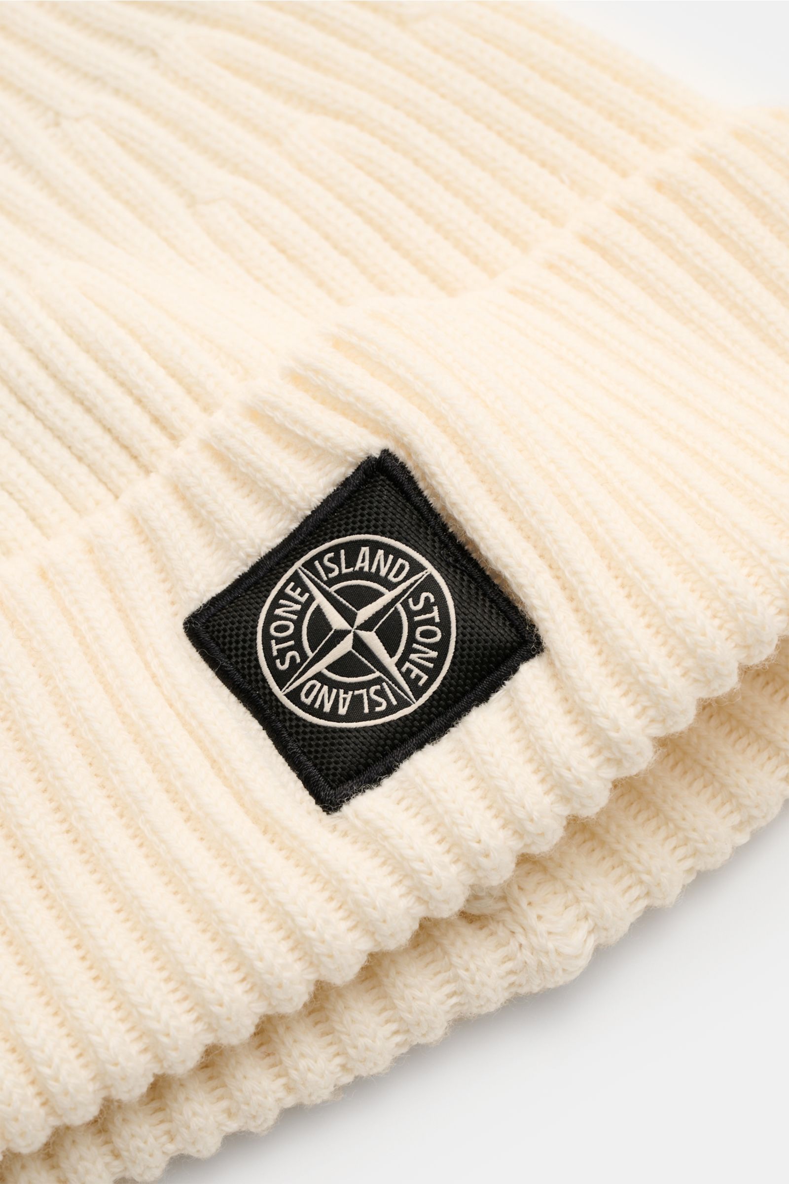 Stone Island Wollmütze creme, Nahaufnahme schräg von oben, reine Wolle, weicher Griff, moderne Rippstrick-Optik, breiter Umschlag mit STONE ISLAND-Weblabel vorne.