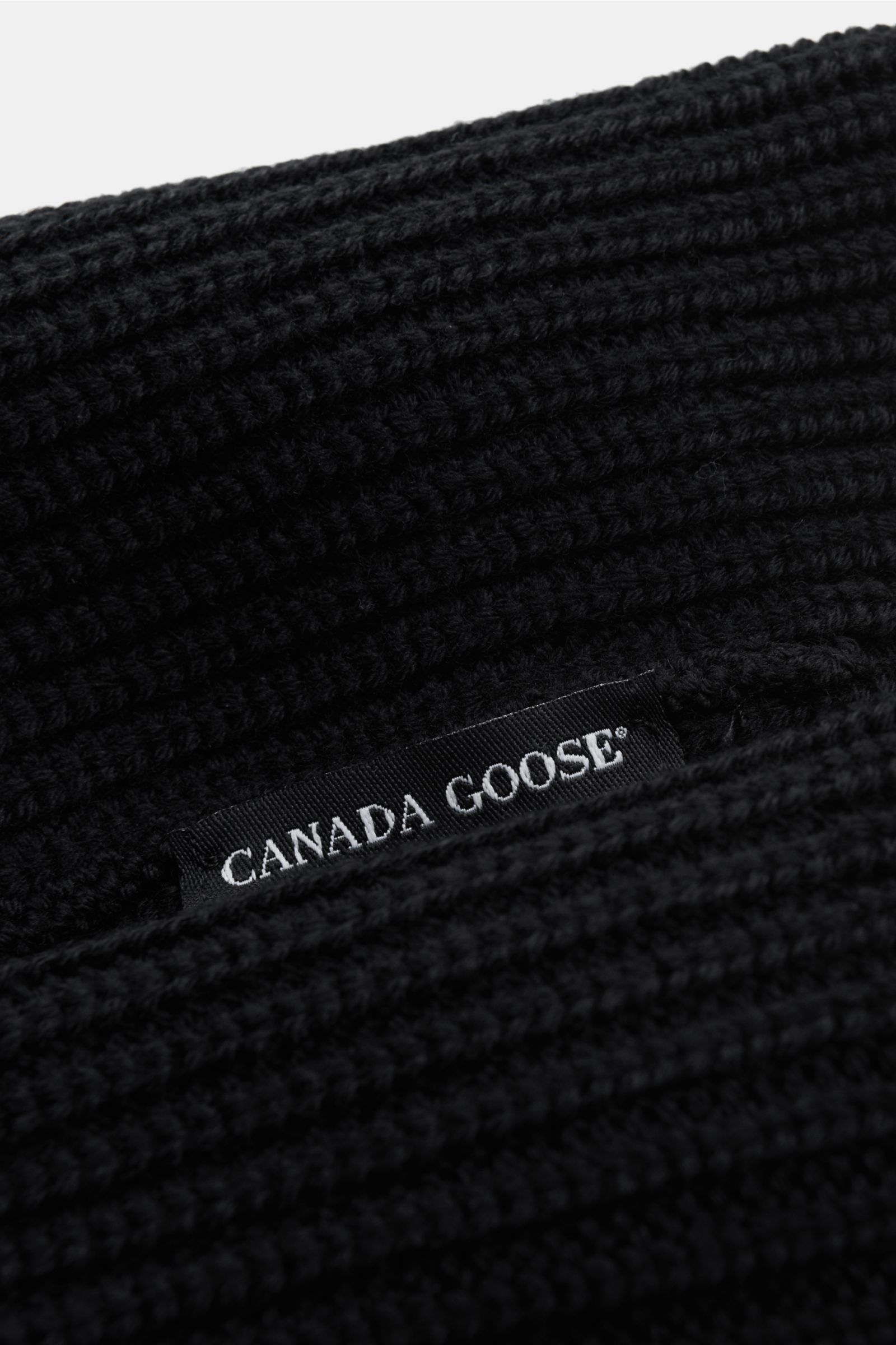 Nahaufnahme aus schräger Perspektive der Canada Goose Wollmütze schwarz aus reiner Wolle mit grobem Rippstrick und Umschlag mit Weblabel.