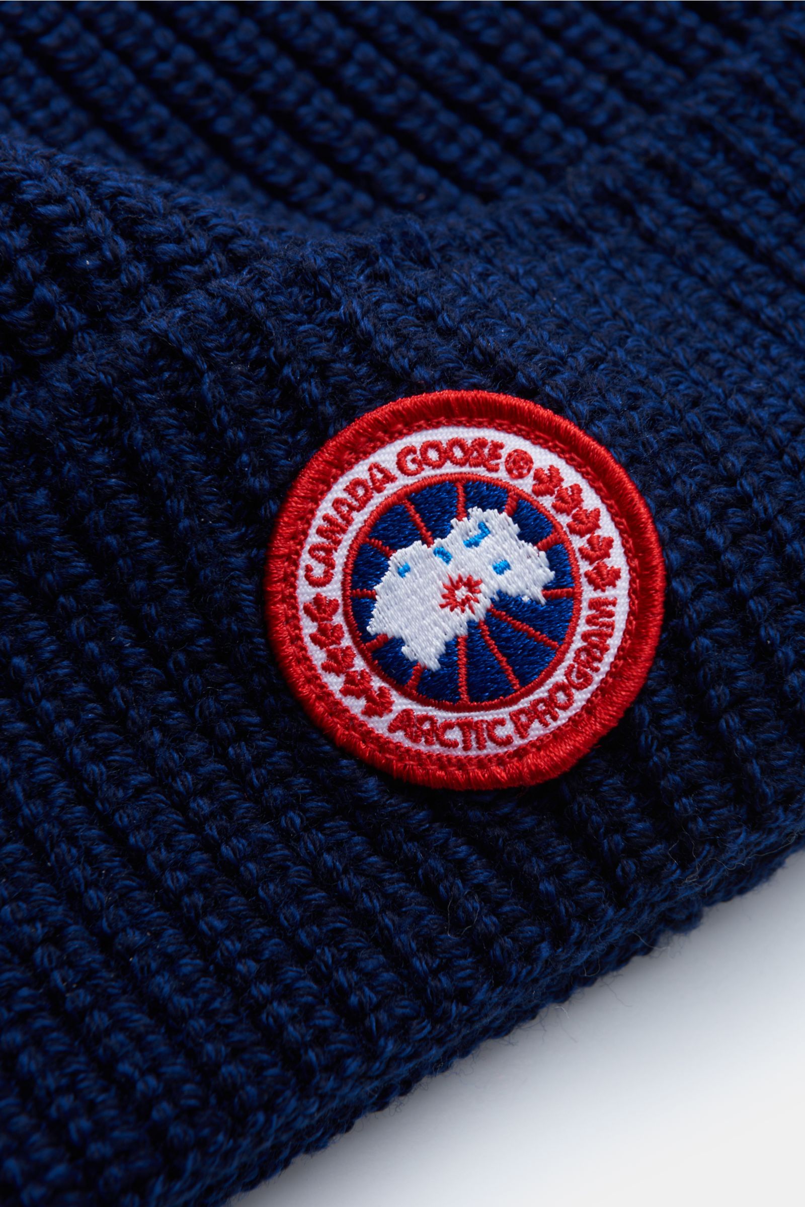 Nahaufnahme der Canada Goose Wollmütze dunkelblau aus grobem Rippstrick mit Umschlag und rotem Weblabel, reine Wolle, griffige Qualität.