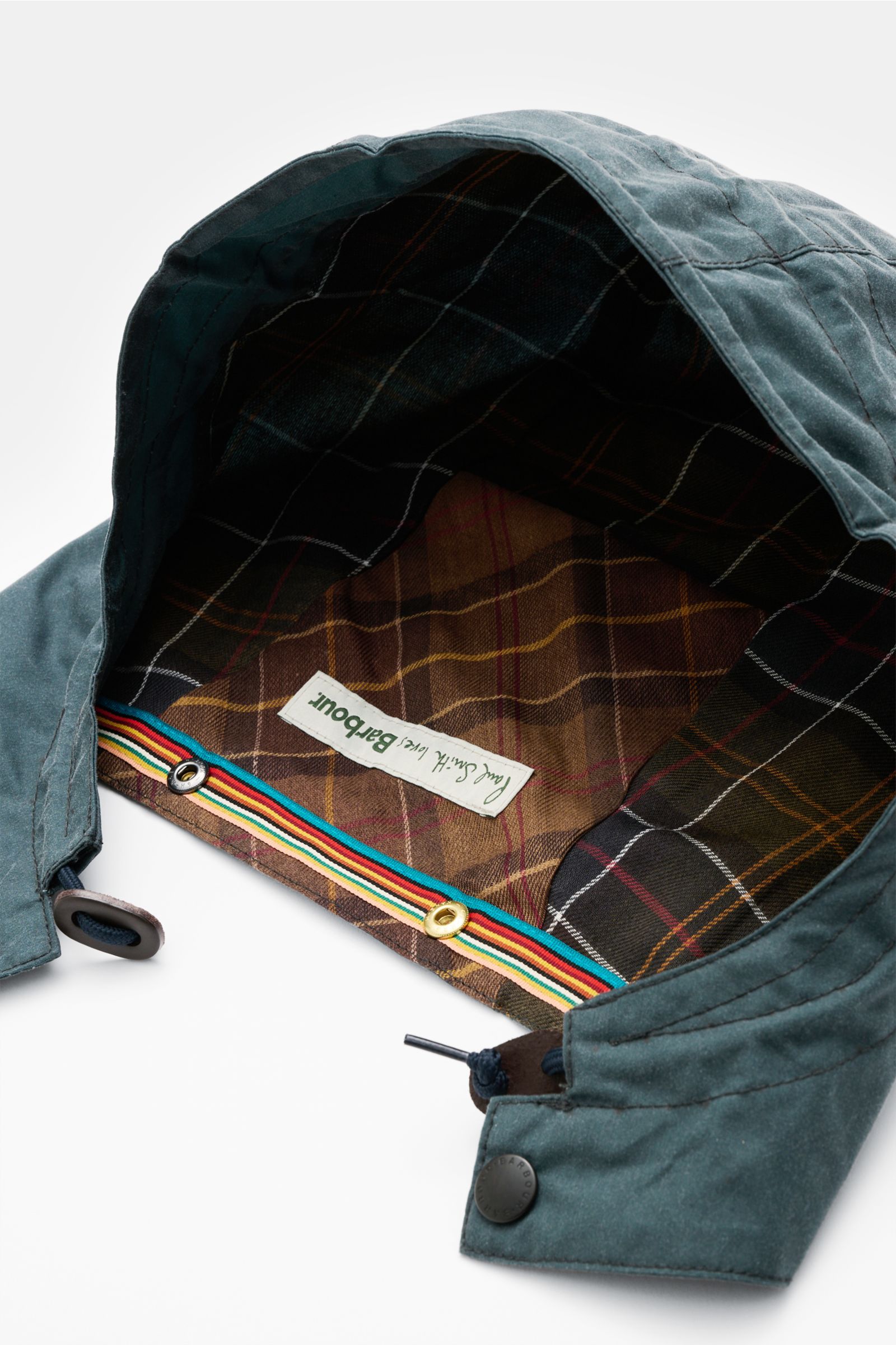 Barbour Kapuze petrol shown from above, highlighting waxed cotton exterior, water-repellent finish, adjustable drawstring, reinforced brim, and signature cotton tartan lining. 

Die Kapuze aus der BARBOUR x PAUL SMITH Kollektion ist mit verschiedenen B