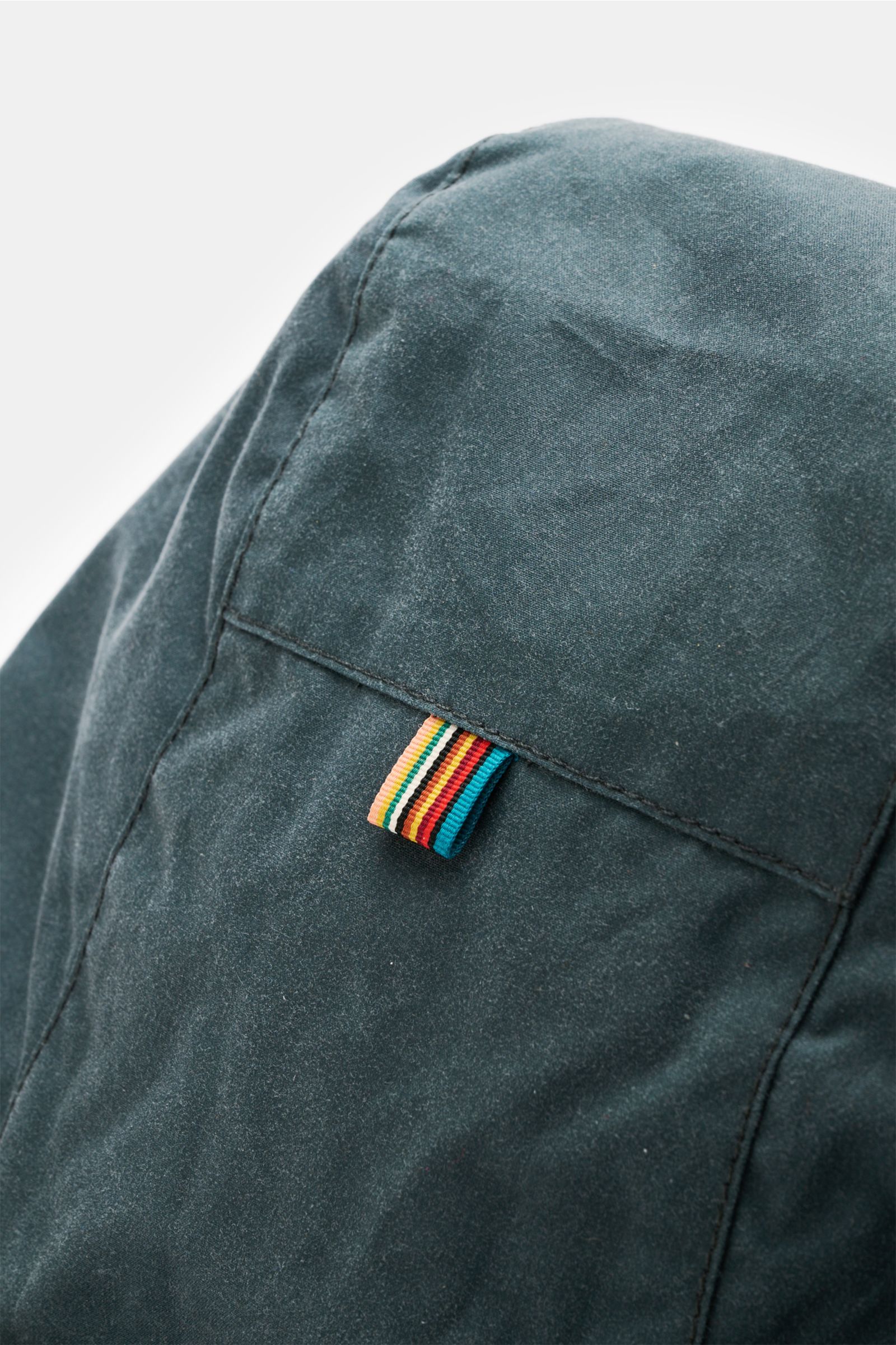 Close-up front view of the Barbour Kapuze petrol hood with waxed cotton fabric, material-typical texture, and colorful striped loop detail.

Die Kapuze aus der BARBOUR x PAUL SMITH Kollektion ist mit verschiedenen BARBOUR-Modellen kompatibel und lässt 