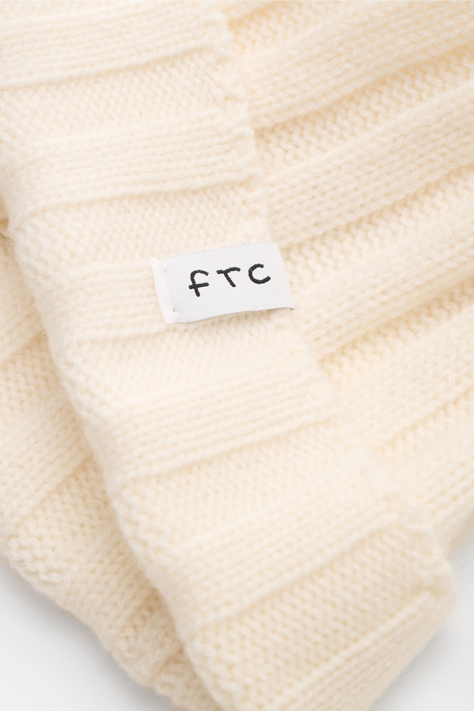 Nahaufnahme der FTC Cashmere Mütze creme aus feinem Fair-Trade-Cashmere mit weichem Griff, Rippstrickmuster und FTC-Weblabel am Umschlag.