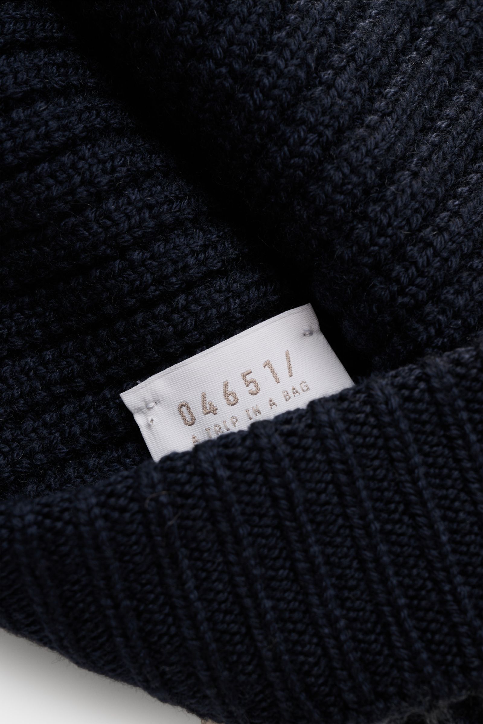 Close-up, angled view of the navy 04651/ A trip in a bag Wool beanie 'Foggy Hat' showing its rib knit pattern and label.