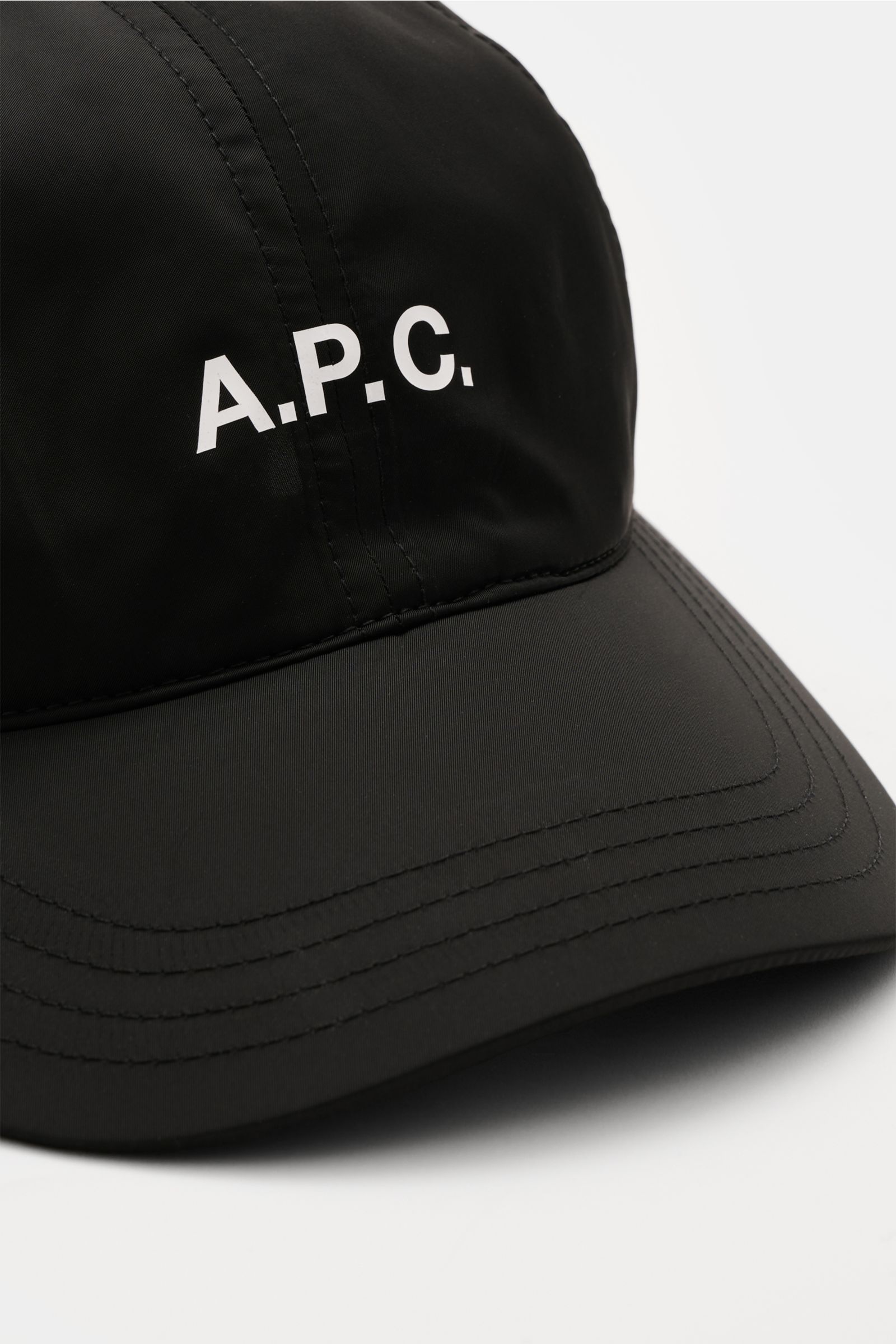 Close-up front-side view of the A.P.C. Baseball-Cap 'Charlie' schwarz in black lightweight microfiber, featuring a classic six-panel design, white logo print on front, and adjustable closure. Lässiger Urban Style aus Paris: Die Baseball-Cap 'Charlie' aus 