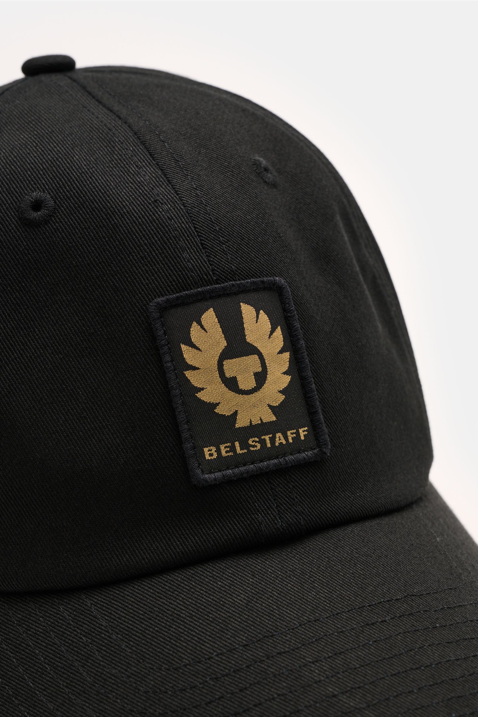 Close-up front-side view of the Belstaff Baseball-Cap schwarz in black cotton twill with robust texture and a gold BELSTAFF woven label on the front. Für sonnige Tage: Die Baseball-Cap aus robustem Baumwolltwill mit Weblabel auf der Front vom Biker-Label 