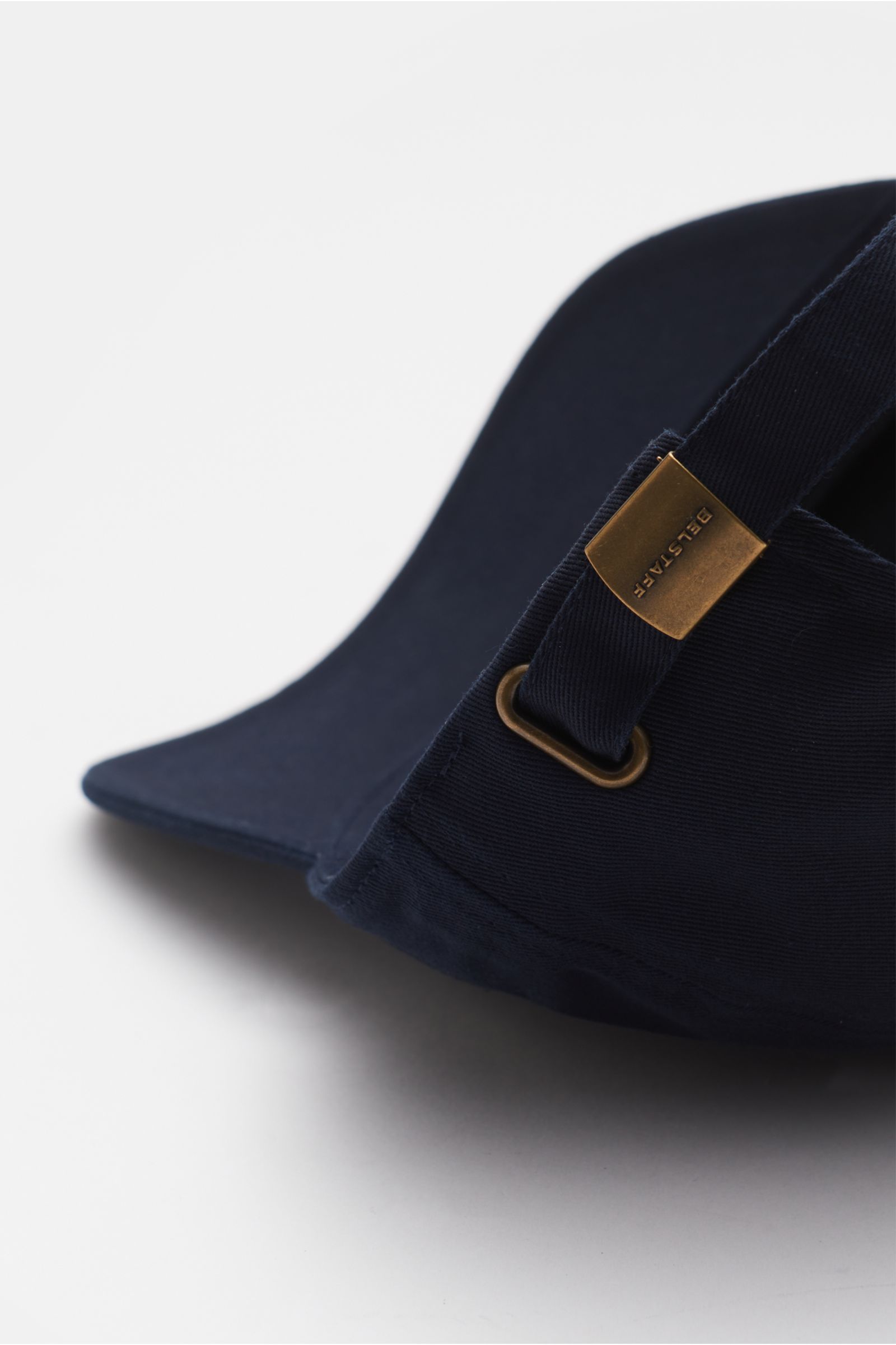 Belstaff Baseball-Cap dark navy shown from a side close-up perspective, made of pure cotton with robust twill texture, featuring an adjustable strap with a brass buckle engraved BELSTAFF and a front web label. Für sonnige Tage: Die Baseball-Cap aus robust