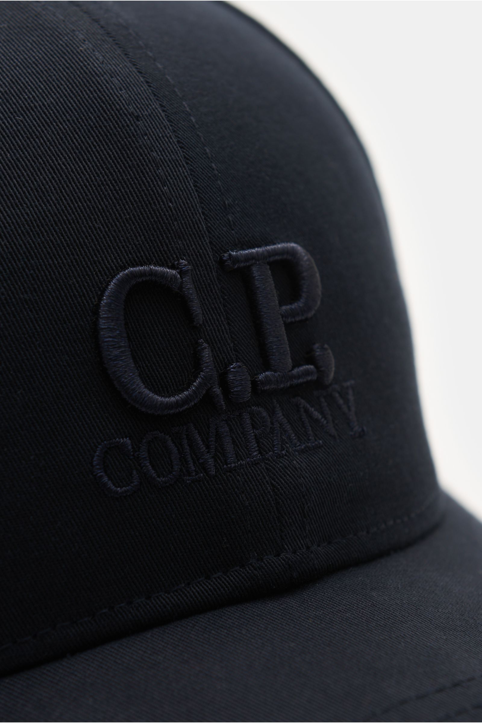 Close-up front-side view of the C.P. Company Baseball-Cap navy, made of pure cotton gabardine with fine weave, lightweight, breathable fabric, garment dyed, featuring a relief-like 3D logo embroidery and an adjustable strap. Die cleane Baseball-Cap aus