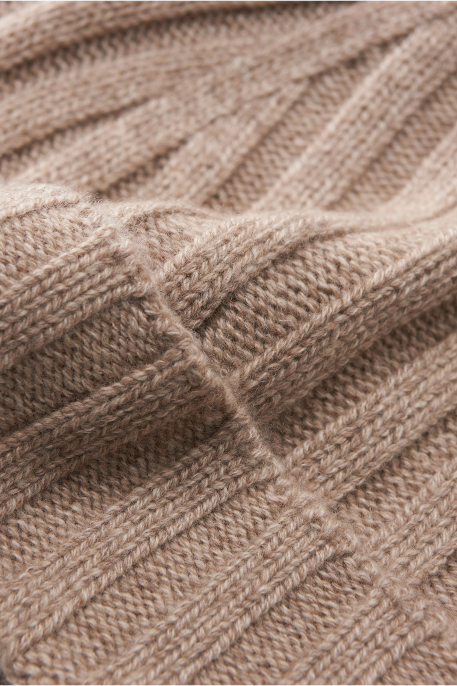 Nahaufnahme der von Braun Cashmere Mütze beige meliert mit klassischem Rippstrickmuster, schmalem Umschlag, feinem Strickbild und sehr weichem Griff aus reinem mongolischem Cashmere, fotografiert aus schräger Perspektive.