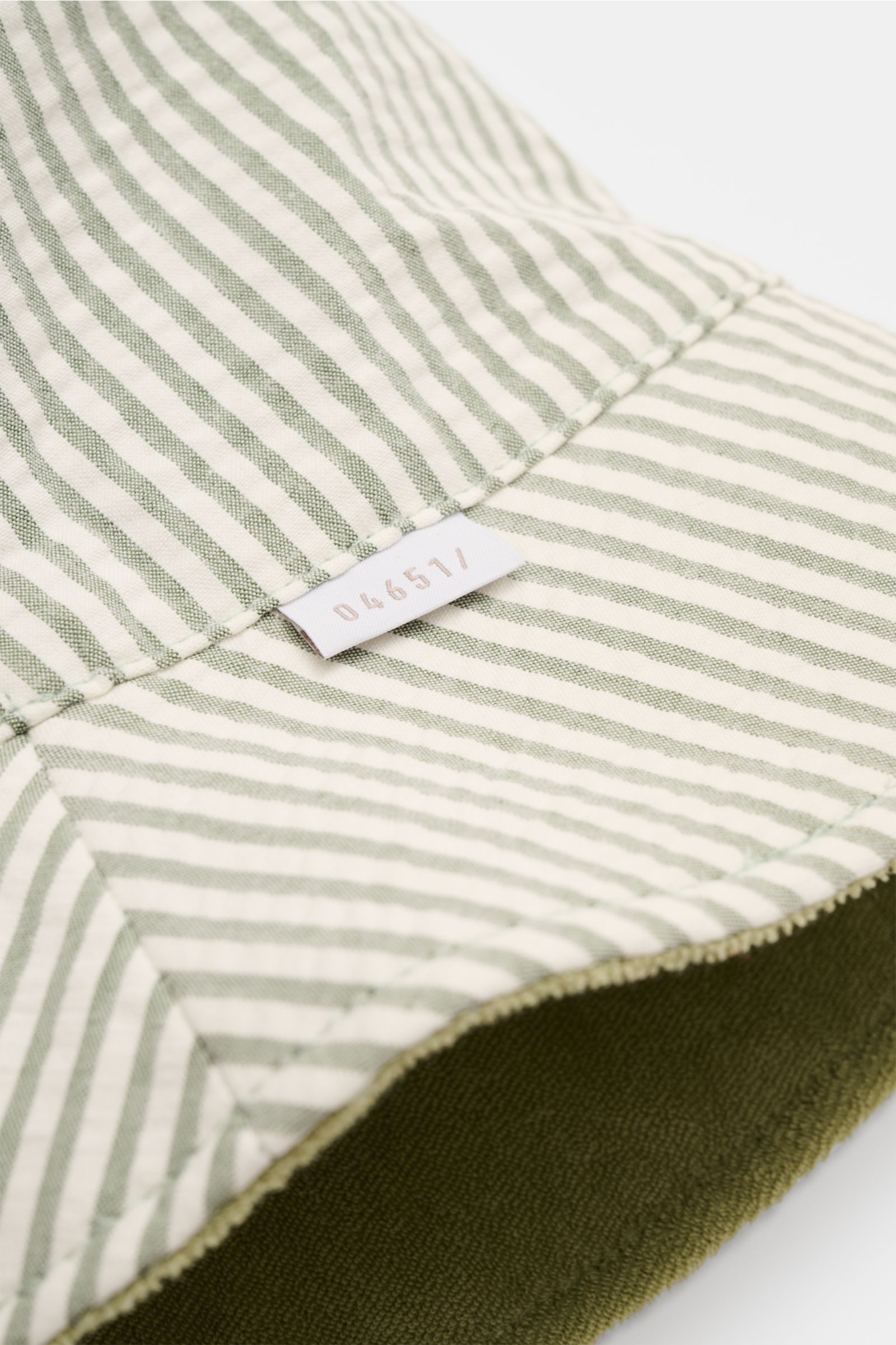Close-up shot from an angled top view of the 04651/ A trip in a bag Wende-Frottee Bucket Hat salbei showing striped seersucker inner lining, soft olive green terry cotton outside, narrow brim, and small 04651 label. Vielseitiger Urlaubsbegleiter: Der Buck