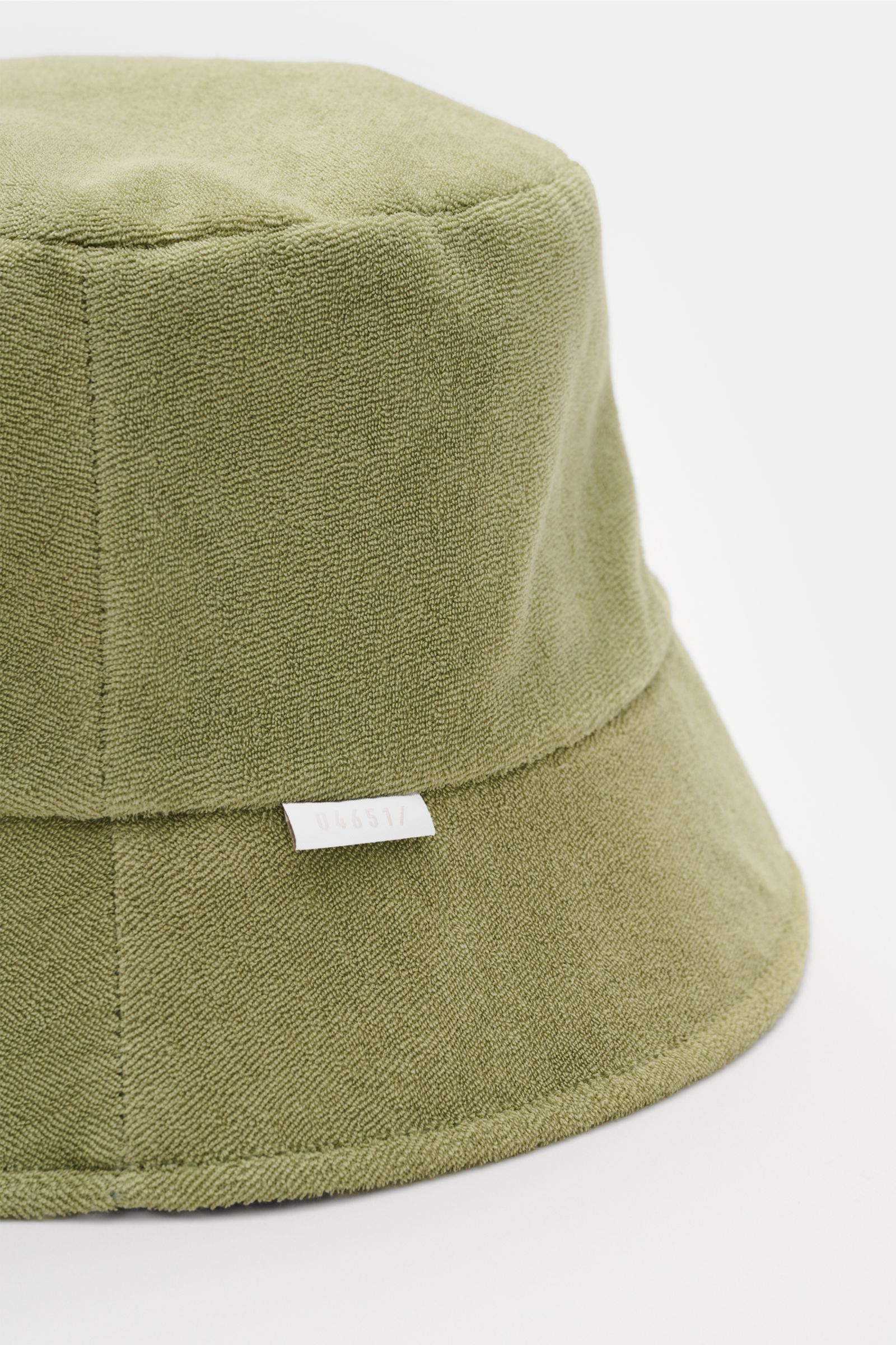 Close-up side view of the 04651/ A trip in a bag Wende-Frottee Bucket Hat salbei in sage green, made from elastic cotton terry with a small label. Vielseitiger Urlaubsbegleiter: Der Bucket Hat von 04651/ A TRIP IN A BAG aus elastischem Baumwoll-Frottee bi