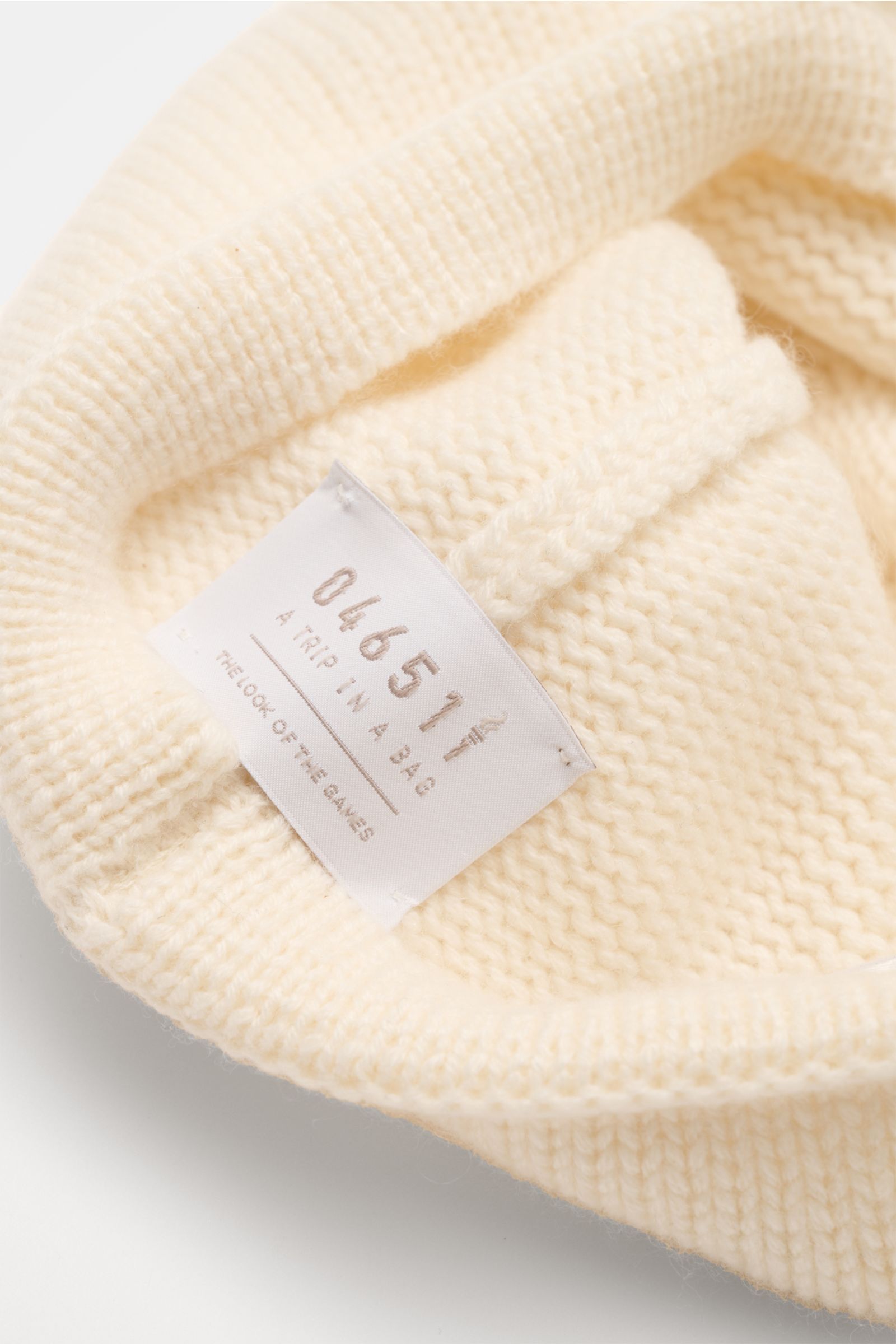 Nahaufnahme der cremefarbenen 04651/ A trip in a bag Cashmere Baseball-Cap aus der 'Look of the Games' Capsule, Detail des reinen Cashmere-Materials mit weichem Griff, mittelgrobem Strickbild und feinstricktem Schirm, Sicht auf das Innenlabel mit kunstvol