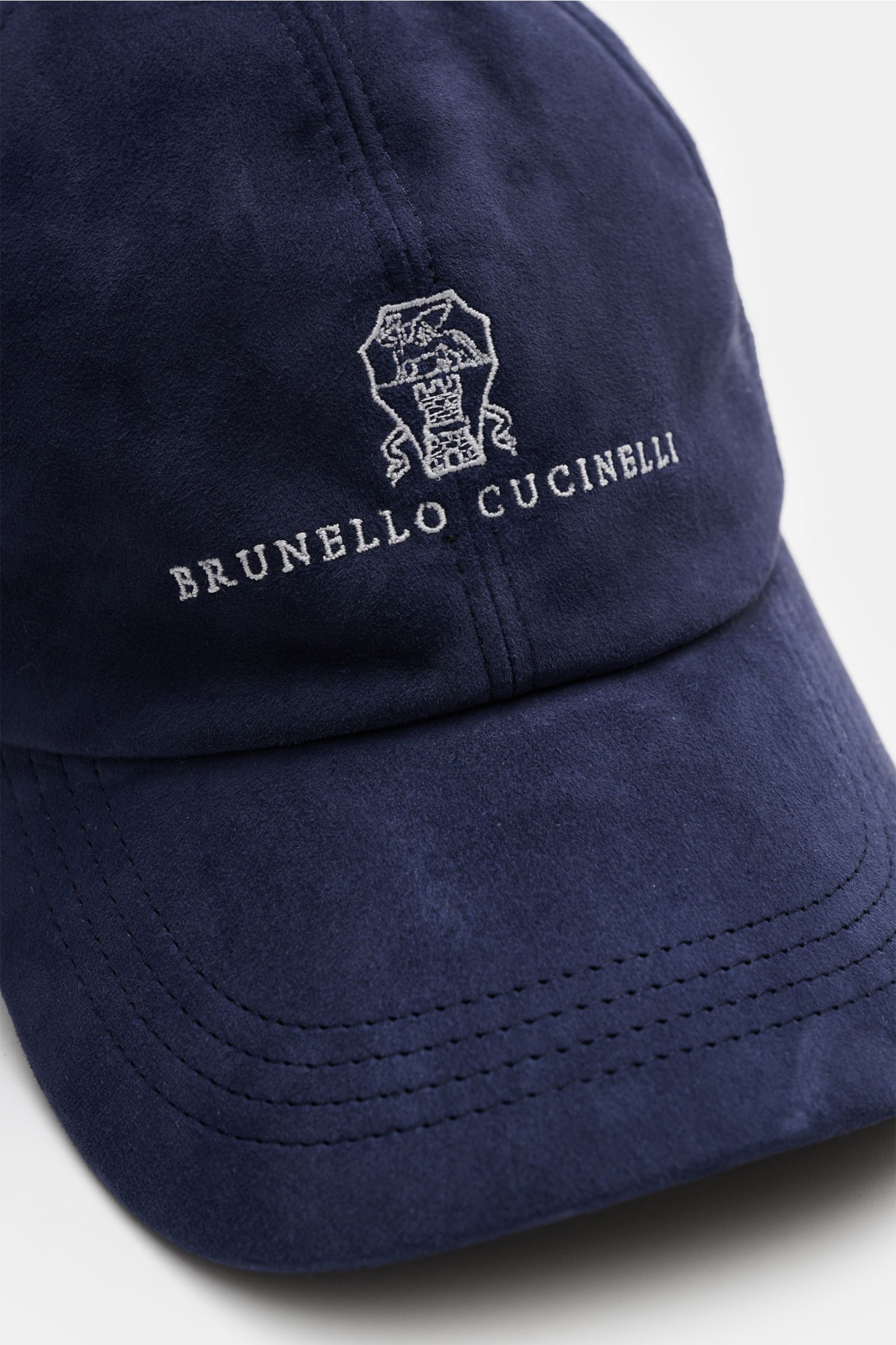 Nahaufnahme der dunkelblauen Brunello Cucinelli Veloursleder-Cap aus softem Veloursleder, klassischer Six-Panel-Stil, Logo-Stitching, leicht gebogener Schirm, verstellbarer Lederverschluss, Futter aus reiner Baumwolle, fotografiert von vorne schräg oben.