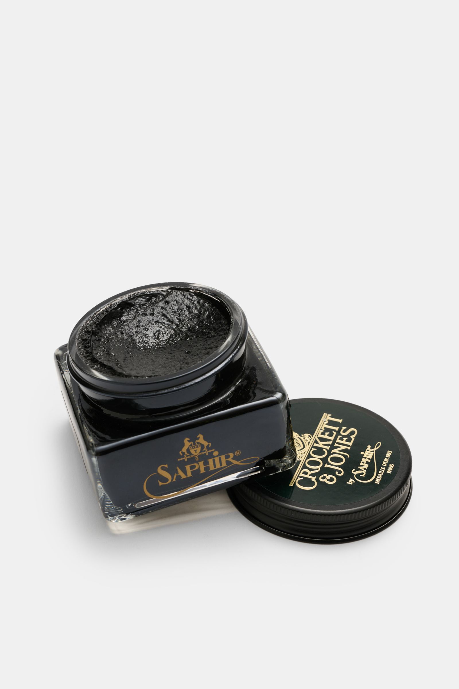 Top-down angled view of Crockett & Jones Shoe polish black 75 ml in a square glass jar with open lid showing black cream polish inside, featuring gold 'Saphir' branding on the jar and 'Crockett & Jones' on the lid. 

A shoe care essential by CROCKETT &