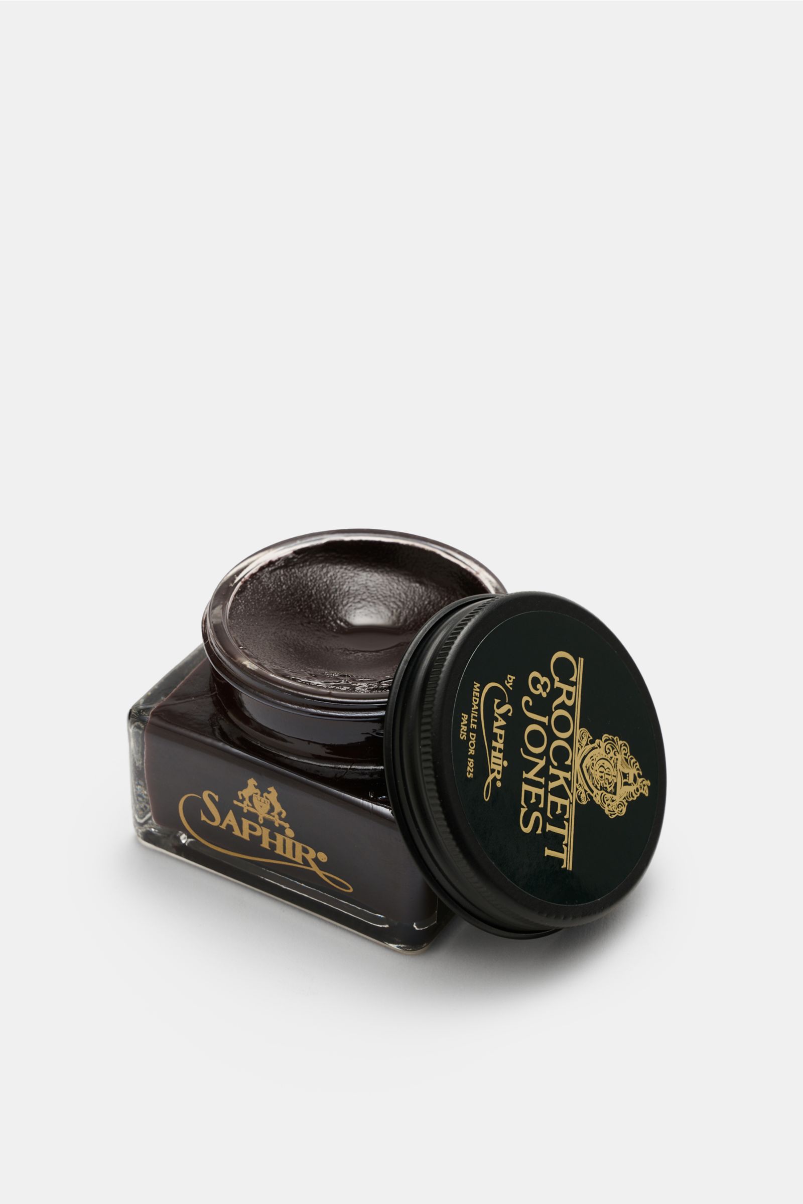 Crockett & Jones Shoe polish brown 75 ml shown from a slightly elevated side angle, open jar with rich dark brown cream polish visible, black lid with gold logo placed beside. 

A shoe care essential by CROCKETT & JONES: This unique cream shoe polish n