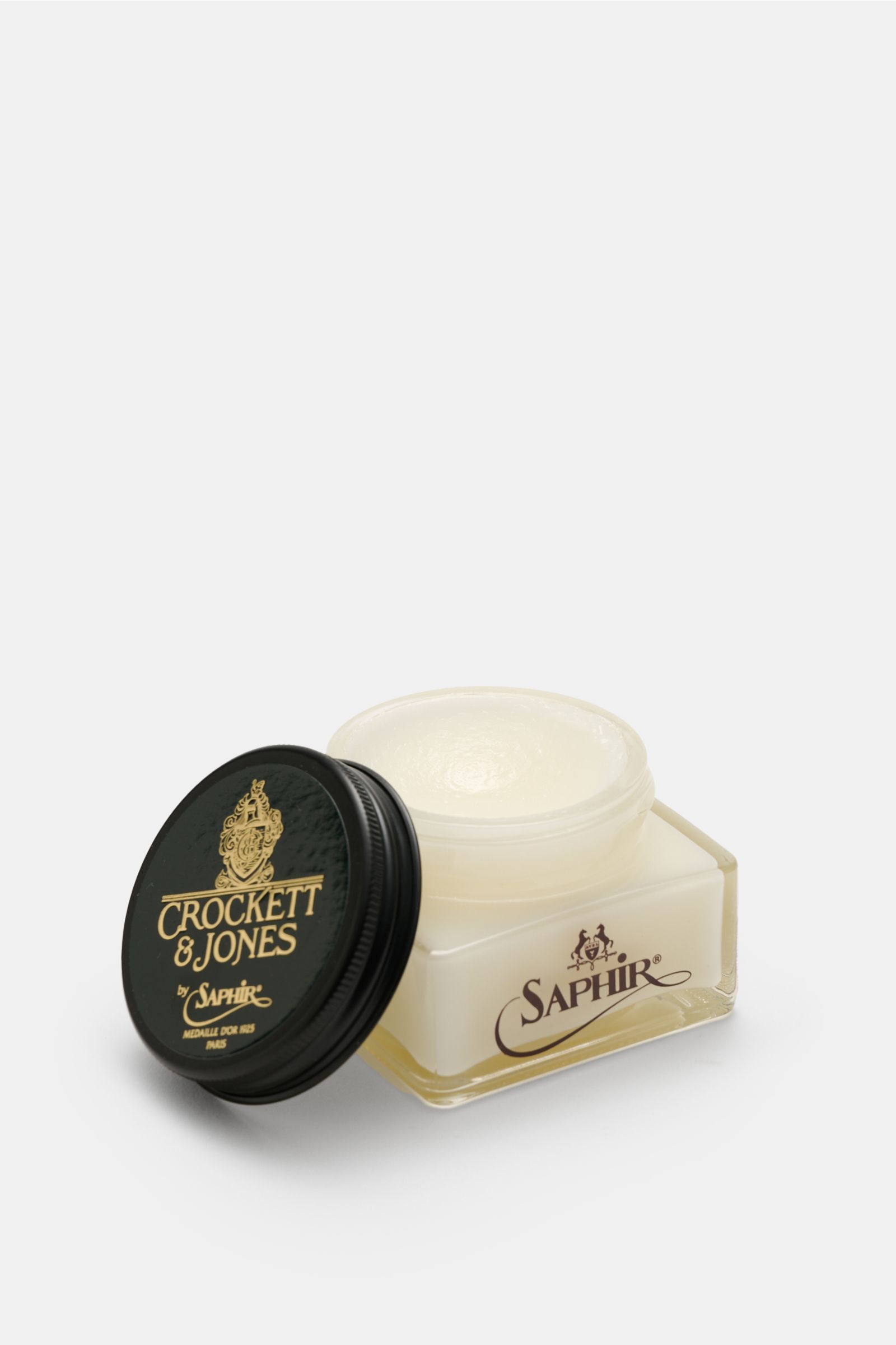 Open jar of Crockett & Jones Shoe polish translucent 75 ml photographed from a front-side angle, showing the creamy polish inside and the black lid with gold branding. 

A shoe care essential by CROCKETT & JONES: This unique cream shoe polish nourishes