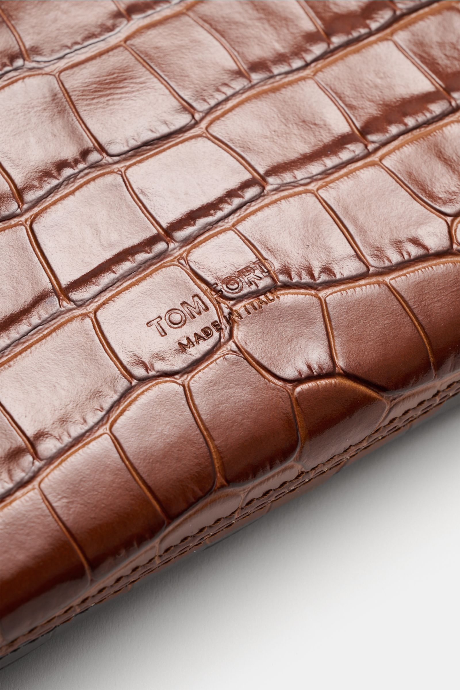 Close-up image of the brown Tom Ford Tasche braun, showing the high-quality calf leather in reptile look with a subtle embossed logo on the front, photographed from a slightly angled top-down perspective. Praktischer Begleiter im Alltag: Hochwertiges Kalb