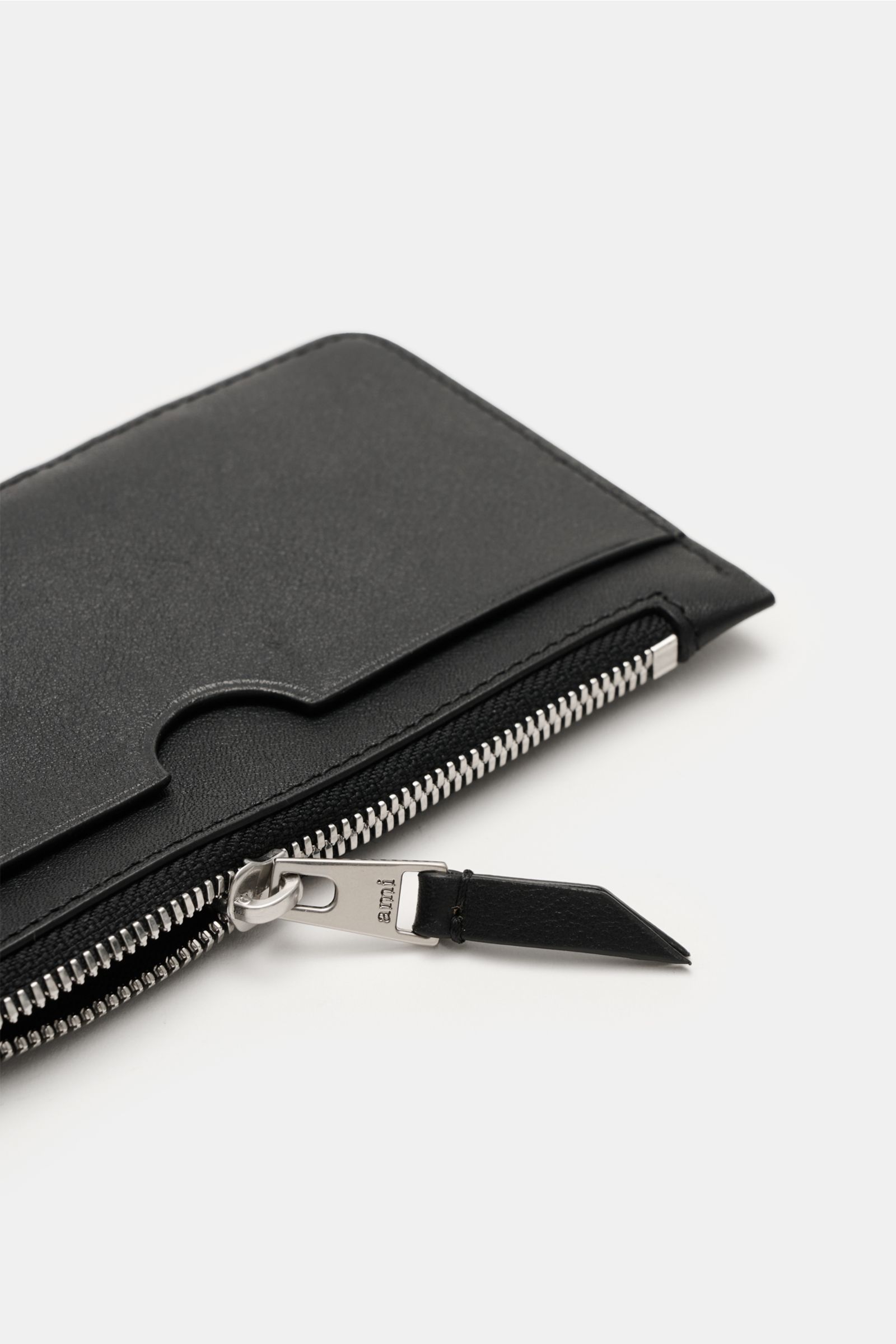 Close-up angled view of Ami Paris Kartenetui schwarz, a black fine-grain calf leather cardholder with a front logo, zippered middle compartment, five card slots, and a large back pocket, measuring approx. 14x8 cm.  
Description:  
Stilvolles Accessoire