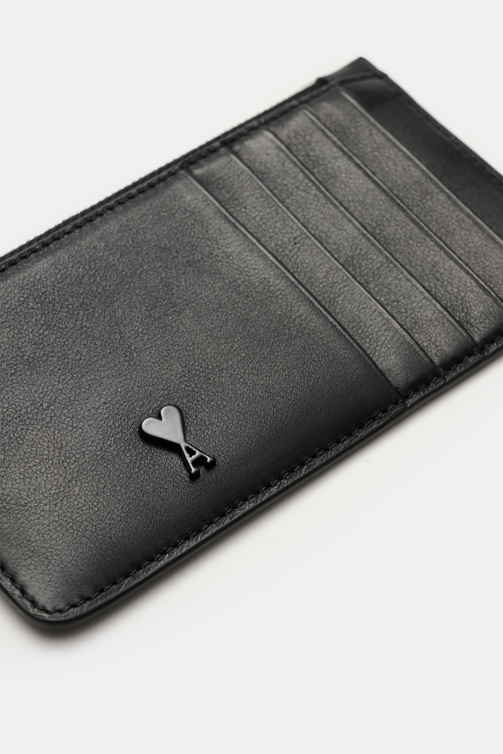 Ami Paris Kartenetui schwarz shown from a slightly angled top-down view, featuring finely grained black calf leather, five card slots, a central zip compartment, a large back pocket, and a front logo heart-shaped 'A'. Stilvolles Accessoire für den Allt