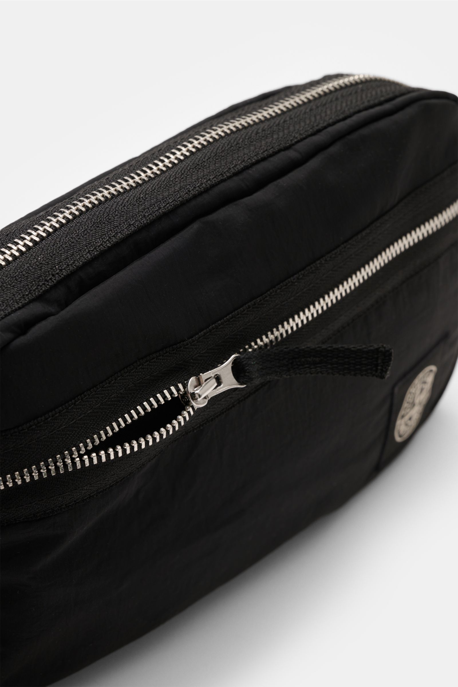 Close-up angled view of Stone Island Gürteltasche schwarz, a minimalist black nylon belt bag with silver zippers, robust strap, front zip pocket, and iconic Windrose patch, approx. 24x15x4.5 cm. Practical Stone Island accessory for everyday essentials lik