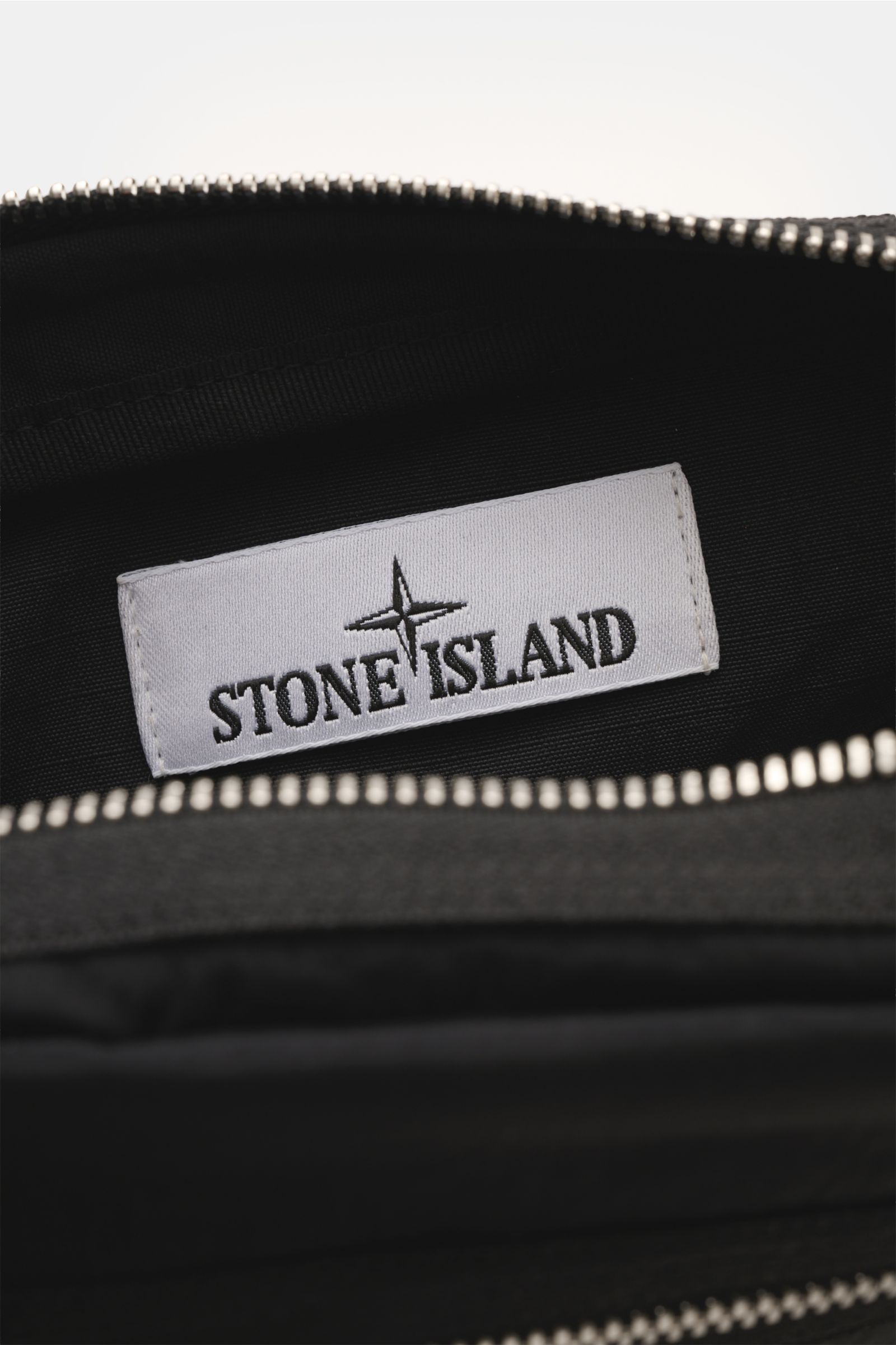 Close-up interior view of the Stone Island Gürteltasche schwarz showing the white Stone Island label inside the black nylon bag with silver zipper teeth.