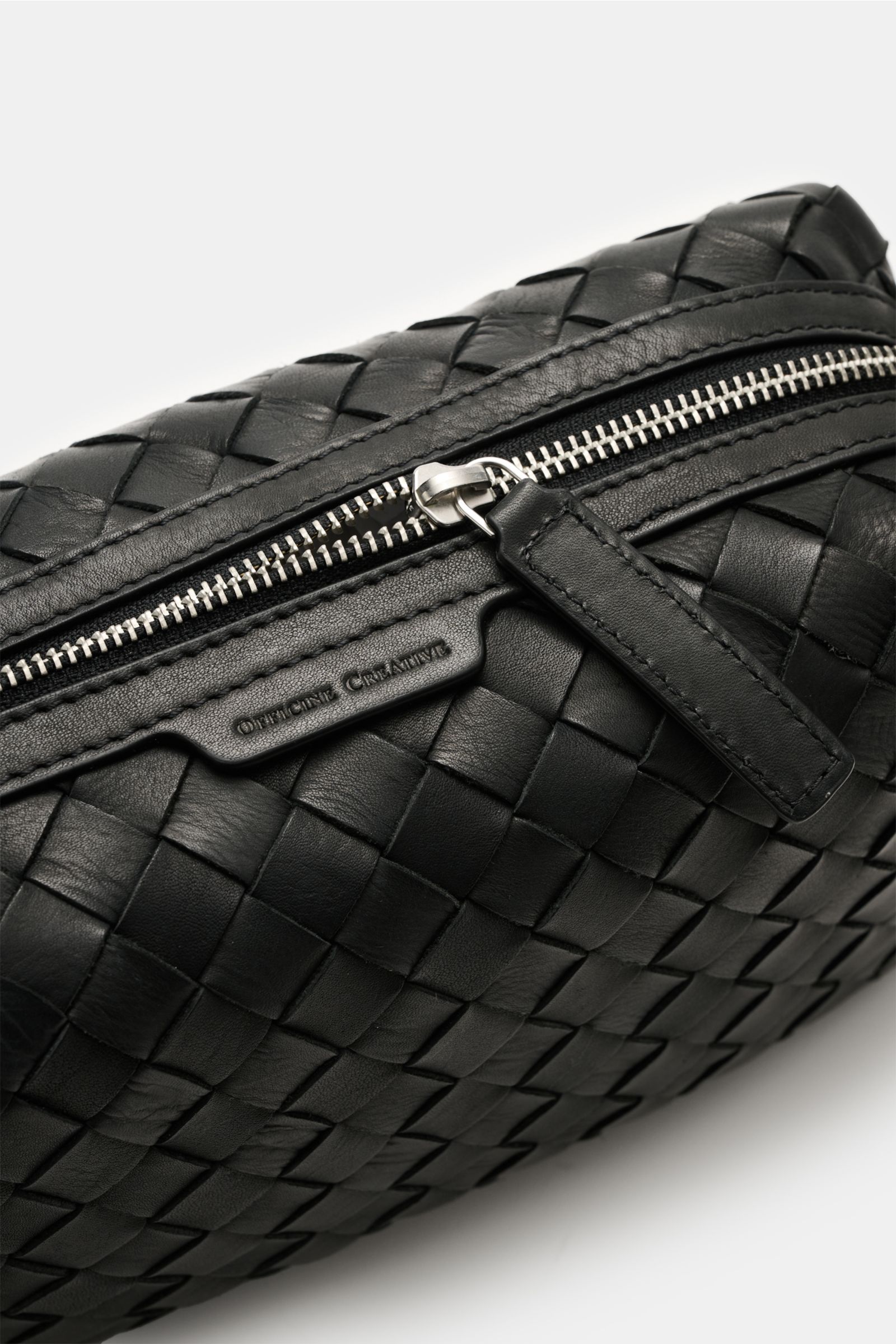 Close-up angled view of the Officine Creative Necessaire schwarz, showing its black woven leather exterior, silver zipper hardware, and embossed logo. Practical travel essential with a spacious main compartment, front zip pocket, and handy wrist strap, si