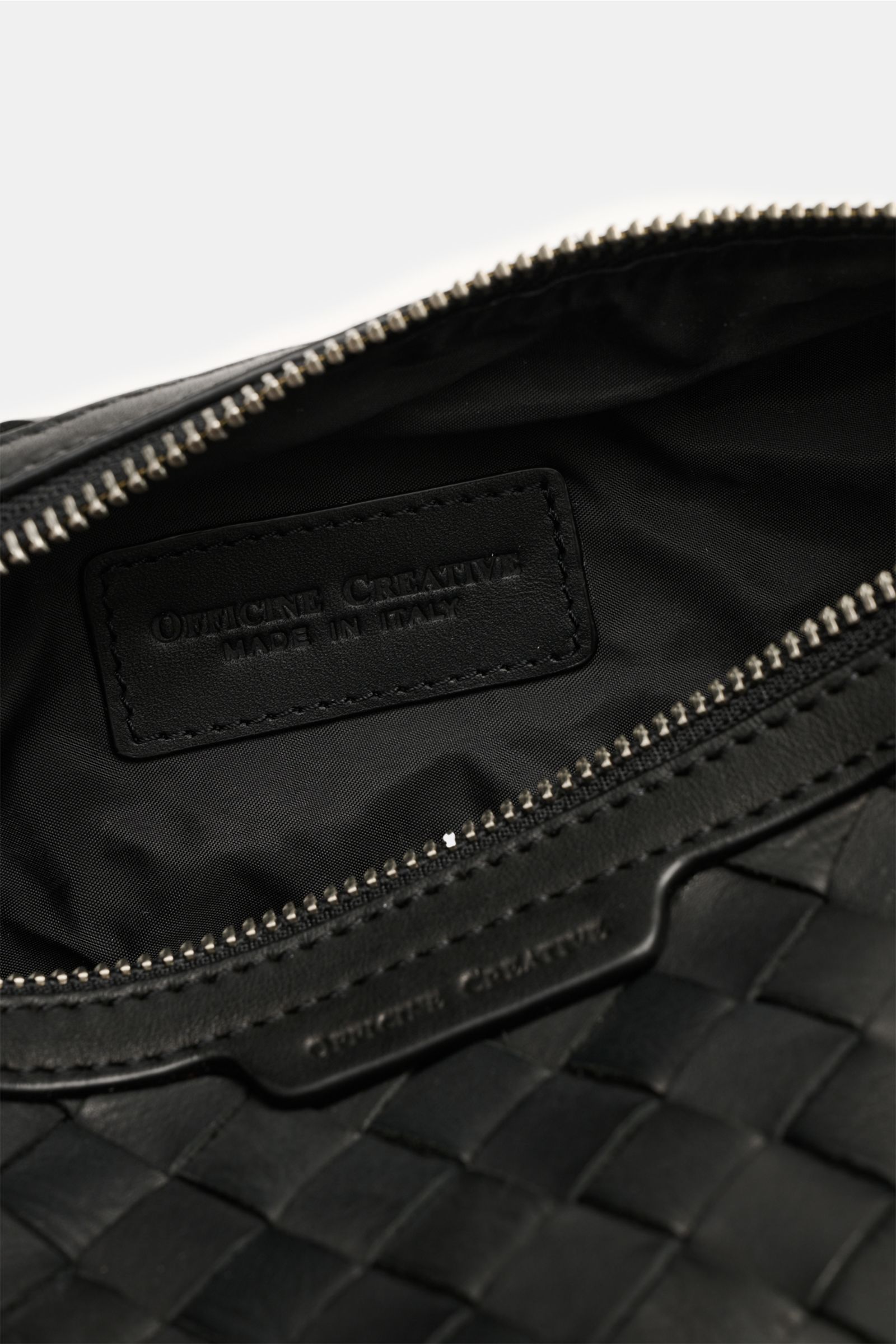 Close-up inside view of the Officine Creative Necessaire schwarz, showing its woven black leather exterior, silver zipper, and stitched brand label. Practical travel essential with spacious main compartment, zipper pocket outside, and handy wrist strap, d