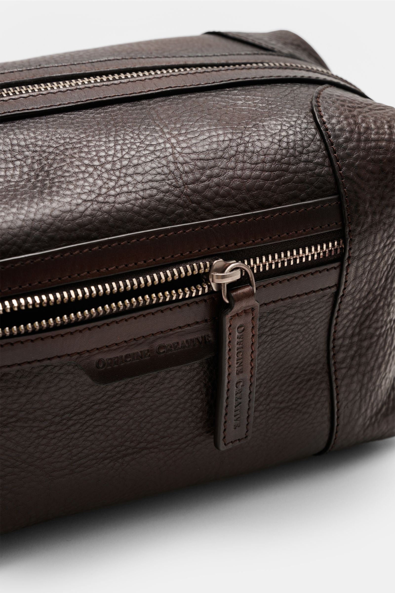 Nahaufnahme des Officine Creative Necessaire dunkelbraun aus fein genarbtem Kalbleder mit silberfarbener Hardware, Frontreißverschluss und Außenfach, seitlich fotografiert.