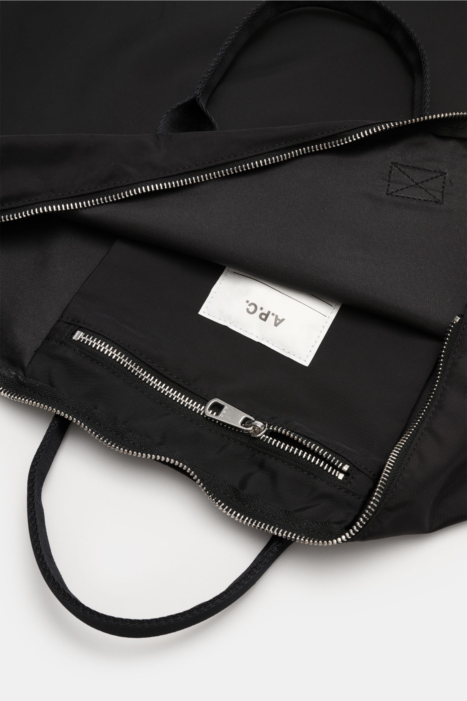 Close-up inside view of A.P.C. Tasche 'Crossbody Bag Backstage' schwarz showing black microfiber, silver zipper, inner zip pocket, and logo label. Versatile everyday companion with durable textile straps, practical shoulder strap, spacious main compartmen