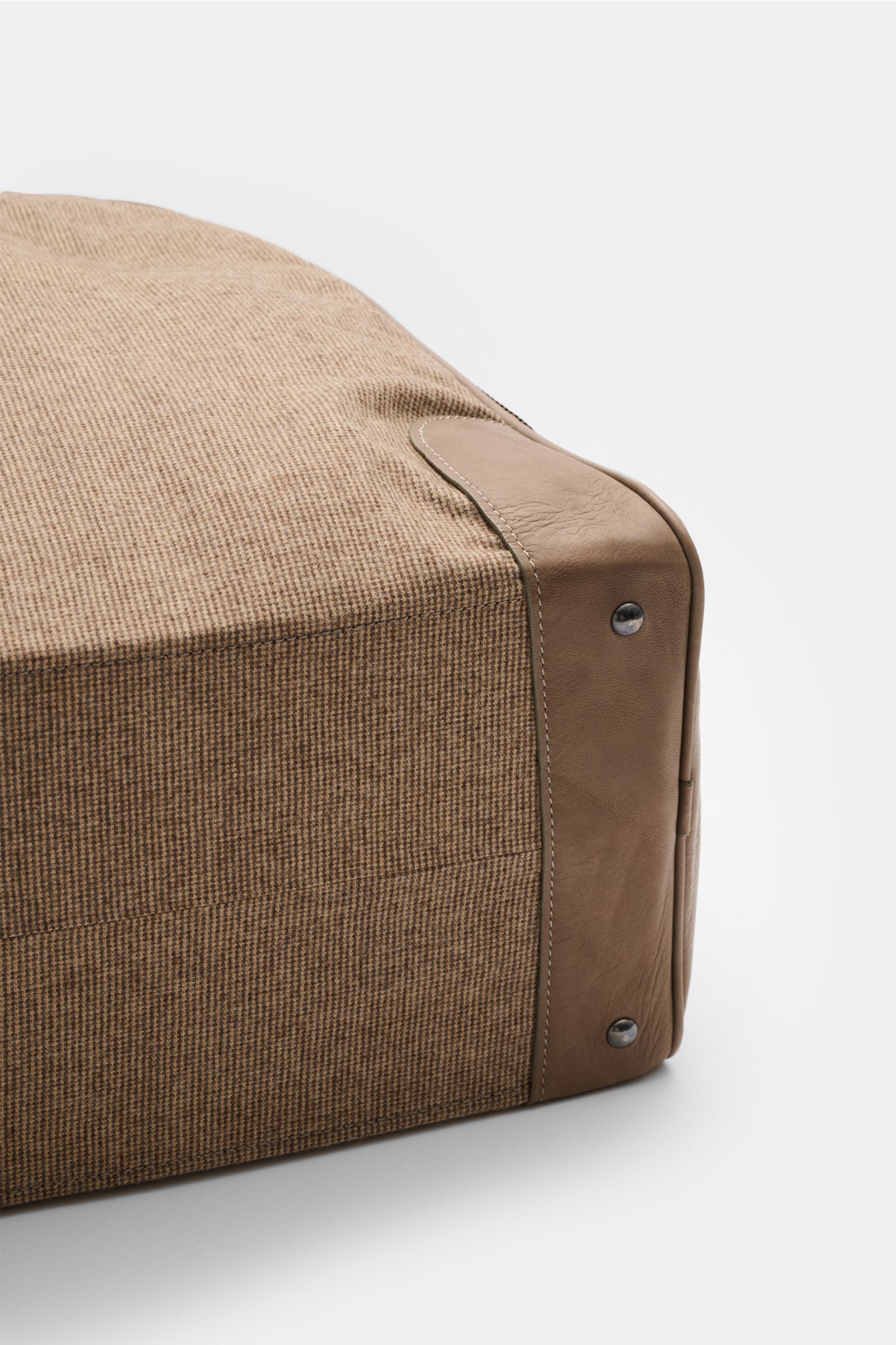 Close-up side view of the bottom corner of the 04651/ A trip in a bag Weekender '356-Bag Tweed' beige/taupe checked, showing fabric and leather with burnished hardware and metal feet. When holiday time comes, you won’t want to be without the weekender '35
