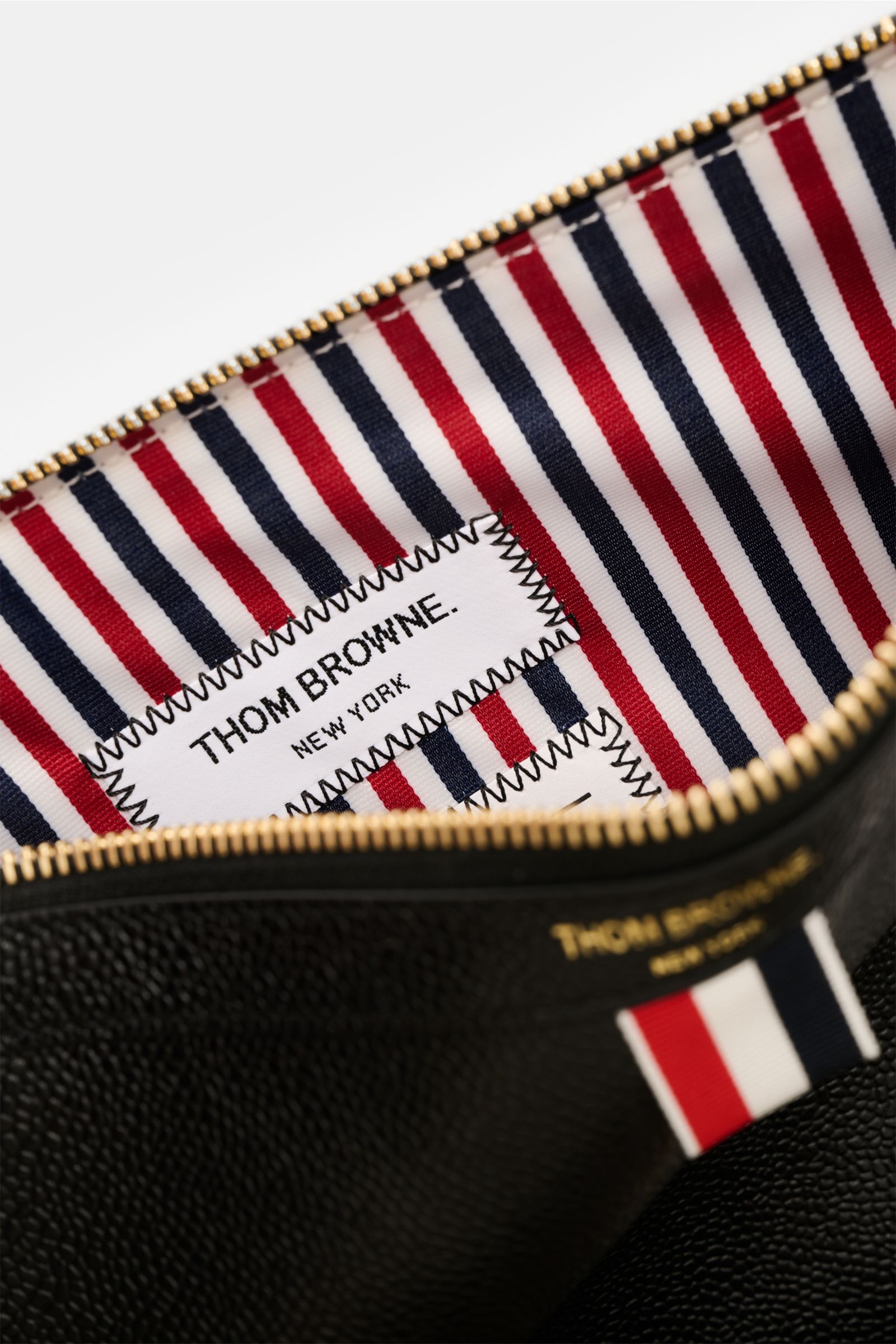 Close-up inside view of the Thom Browne Dokumentenmappe schwarz showing striped interior fabric with a stitched brand label and gold zipper from above.

Die Dokumentenmappe aus edlem Leder von THOM BROWNE wird mit dem markentypischen Grosgrain zum Eyec