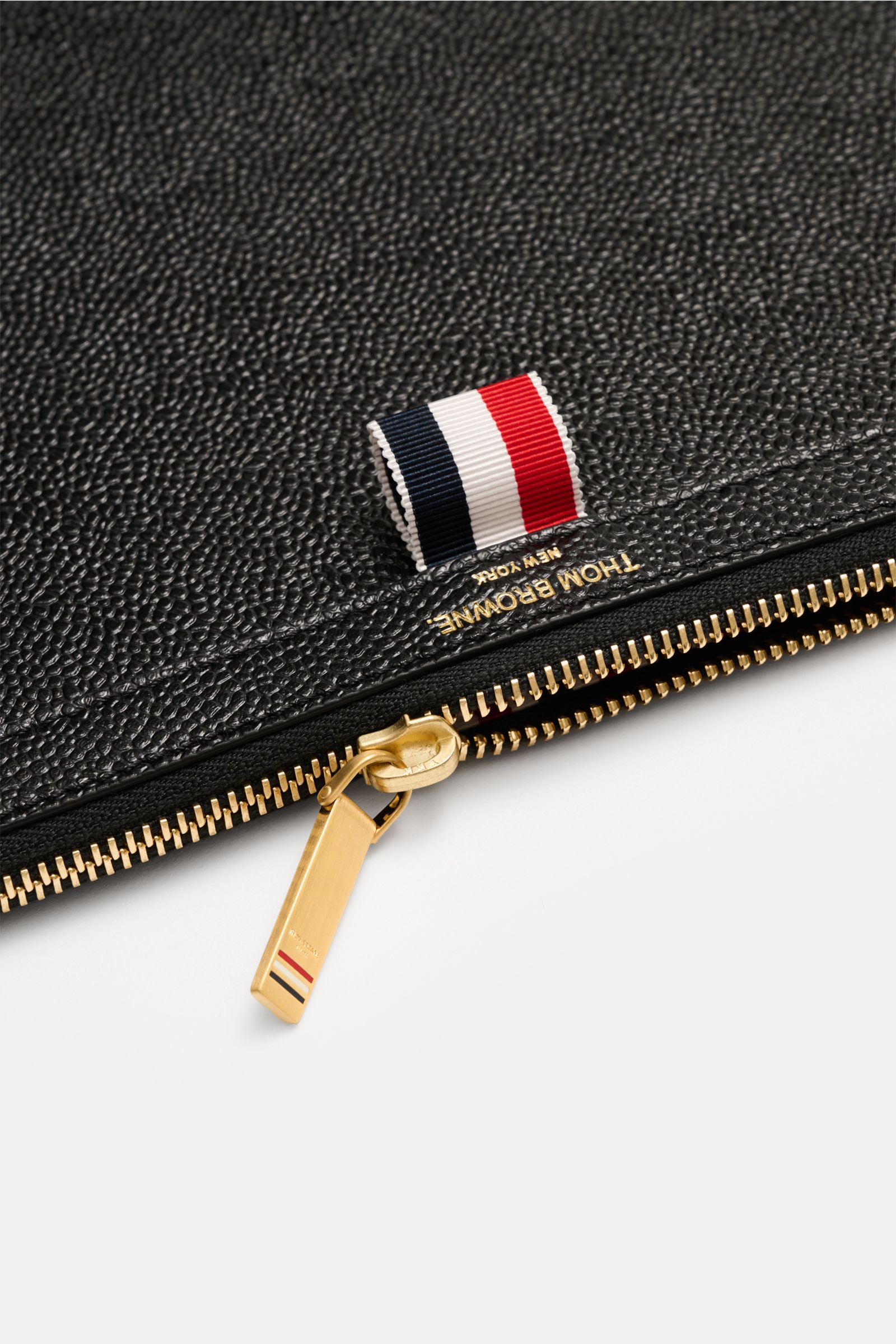 Thom Browne Dokumentenmappe schwarz aus fein genarbtem Leder, goldfarbener Reißverschluss, Grosgrain-Detail, Nahaufnahme von schräger Perspektive.