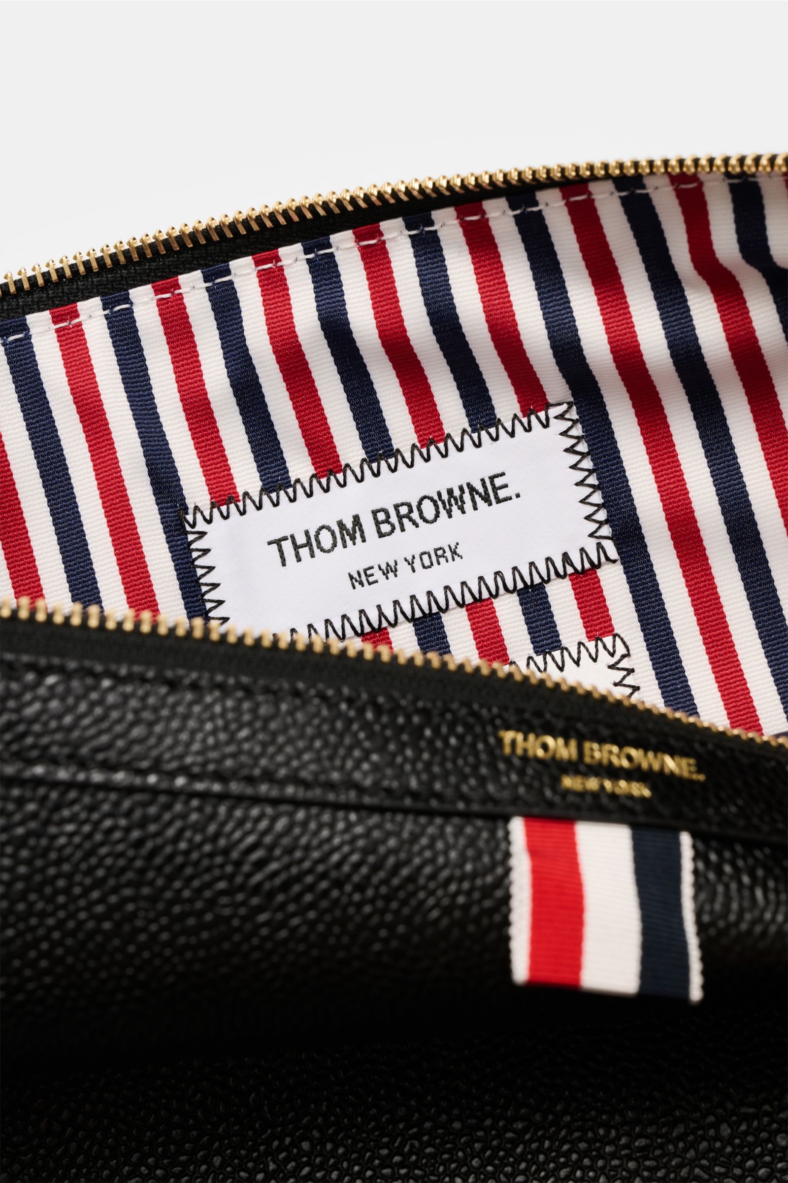 Nahaufnahme der geöffneten Thom Browne Dokumentenmappe schwarz aus fein genarbtem Leder, goldfarbene Hardware, Innenfutter mit rot-blau-weißem Streifendesign, Blick von oben ins geräumige Hauptfach.